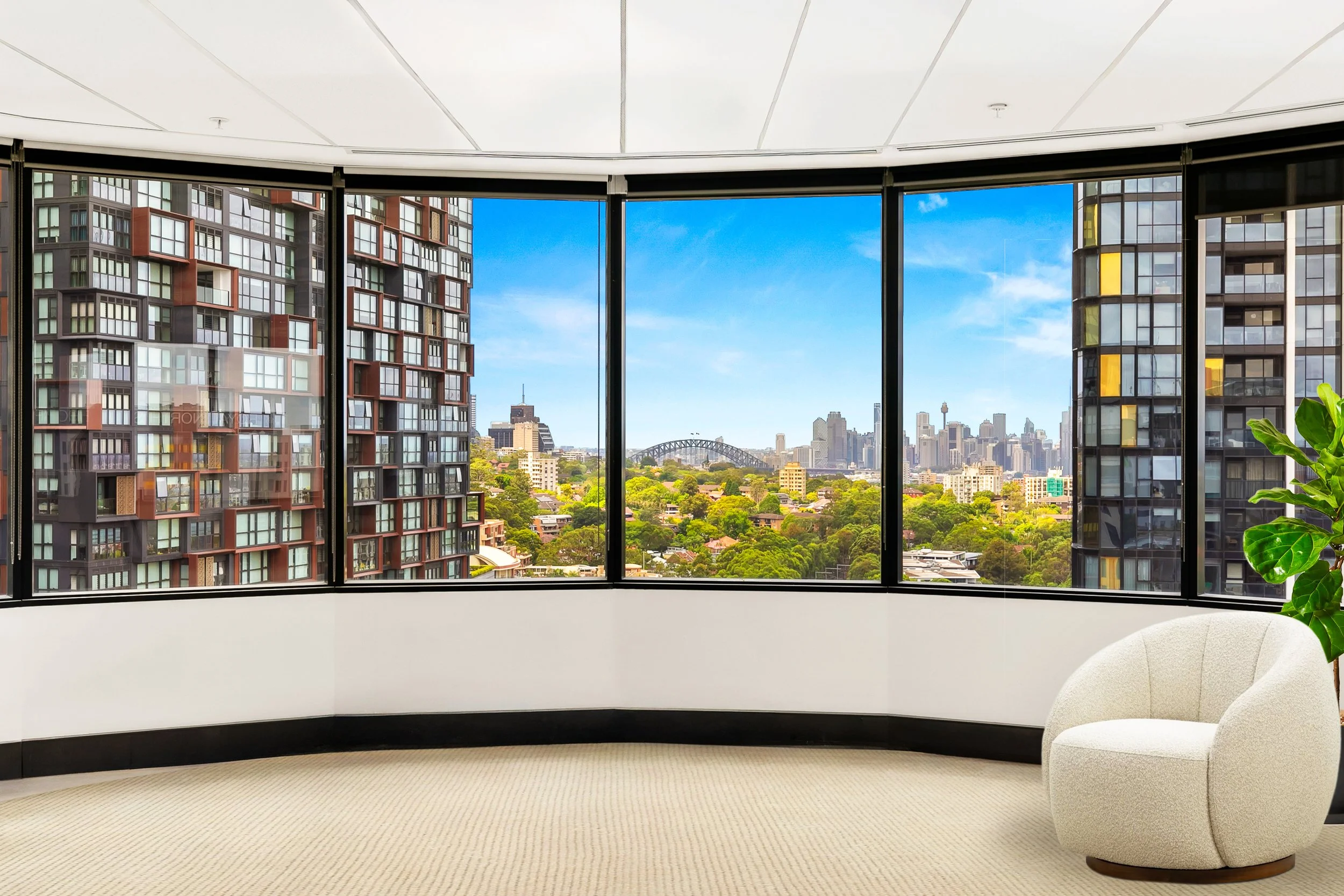 Interior of a high-rise office suite with large curved floor-to-ceiling windows showing a Sydney skyline within St Leonards. There is a white modern chair and a potted green plant in the room.