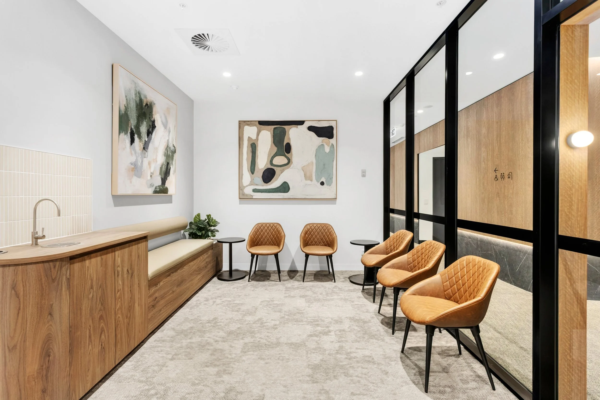 Modern waiting area with six tan leather chairs, black side tables, a built-in wooden counter with a sink, abstract artwork on white walls, a carpeted floor, and glass partitions with wood paneling.