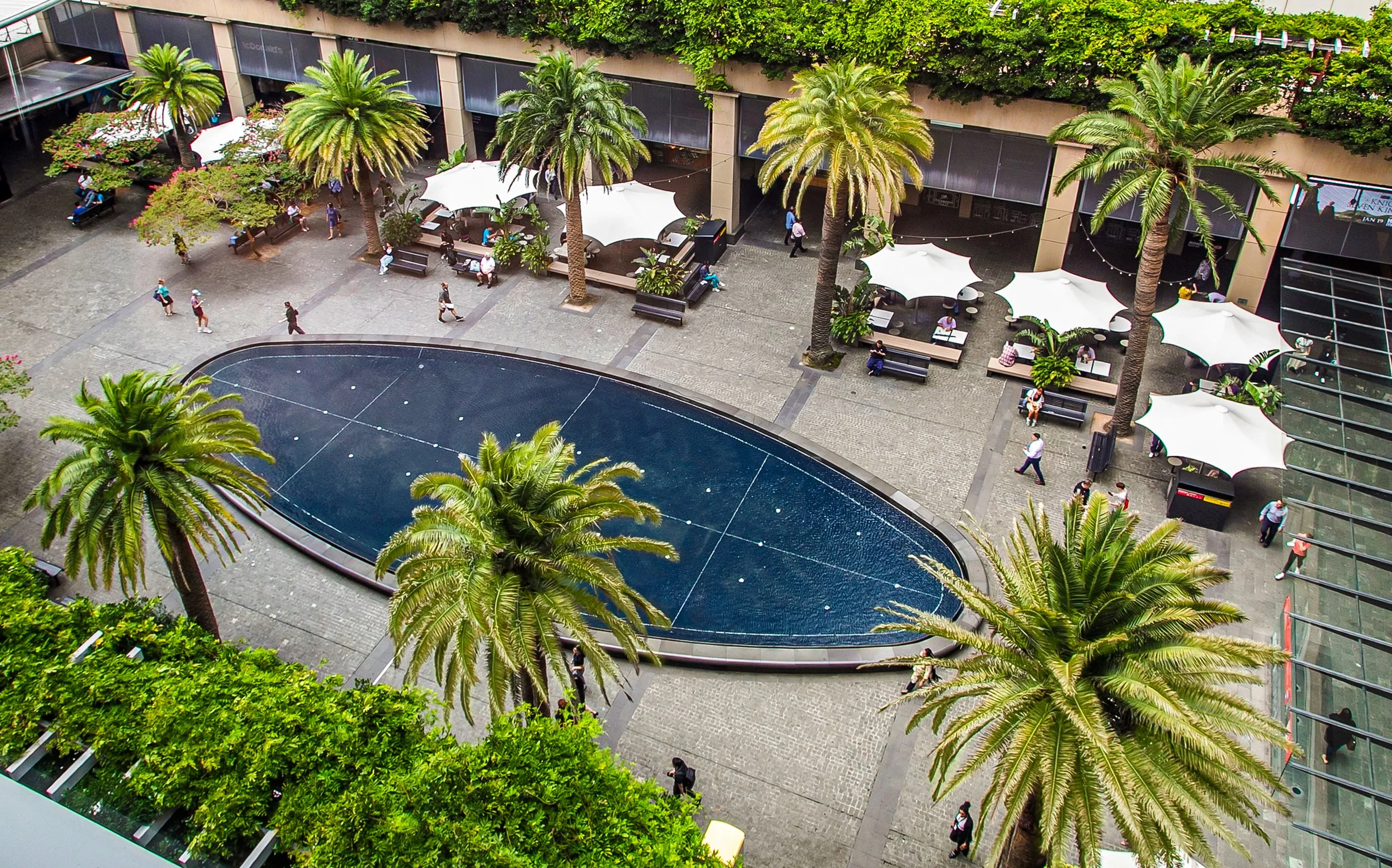 An aerial view of Forum Plaza featuring a large oval pool, tall palm trees, outdoor seating with white umbrellas, and pedestrians walking towards St Leonards Train Station
