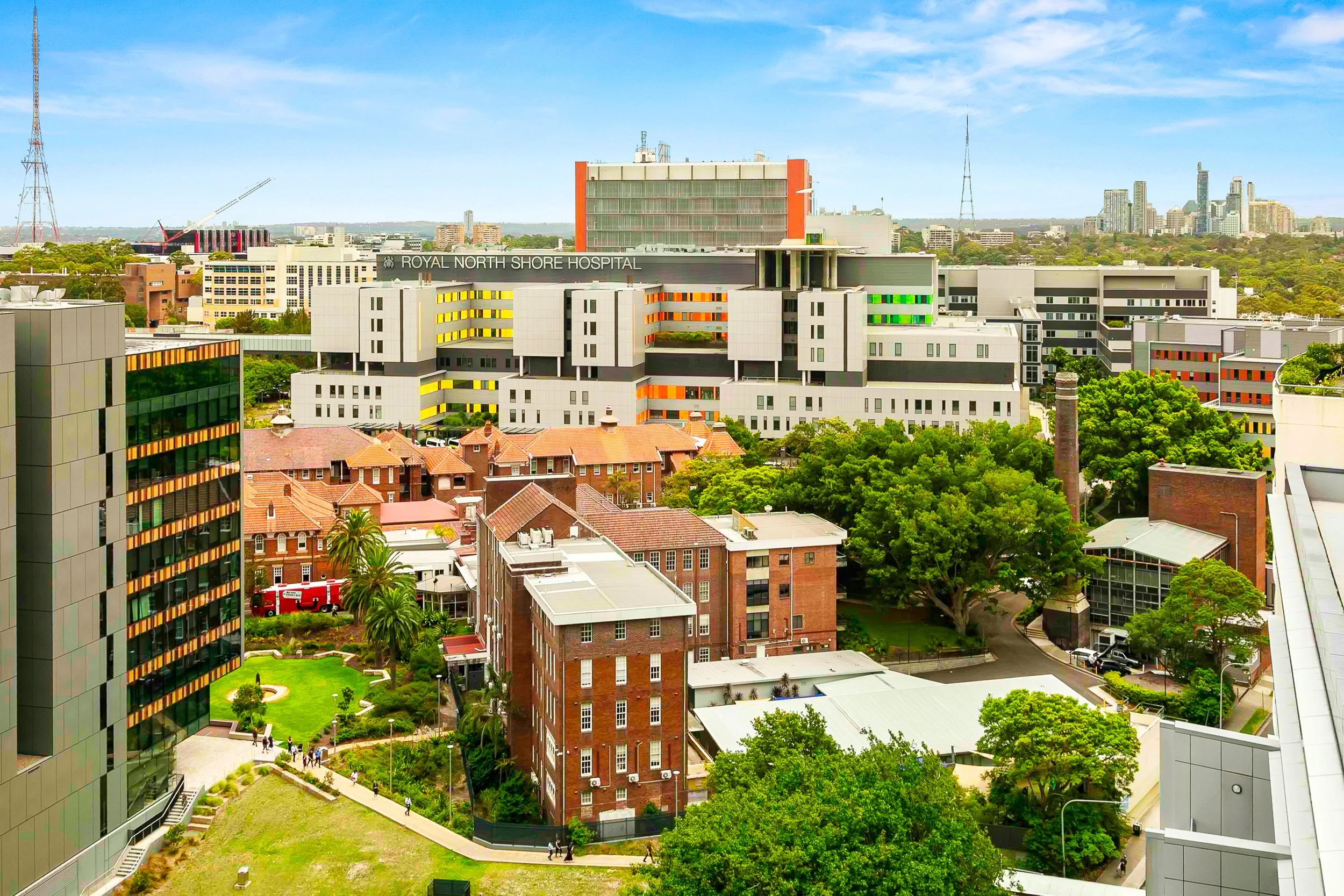 A view with a modern hospital building labeled 'Royal North Shore Hospital' in the background, surrounded by trees, brick buildings, and other modern structures, under a partly cloudy sky.