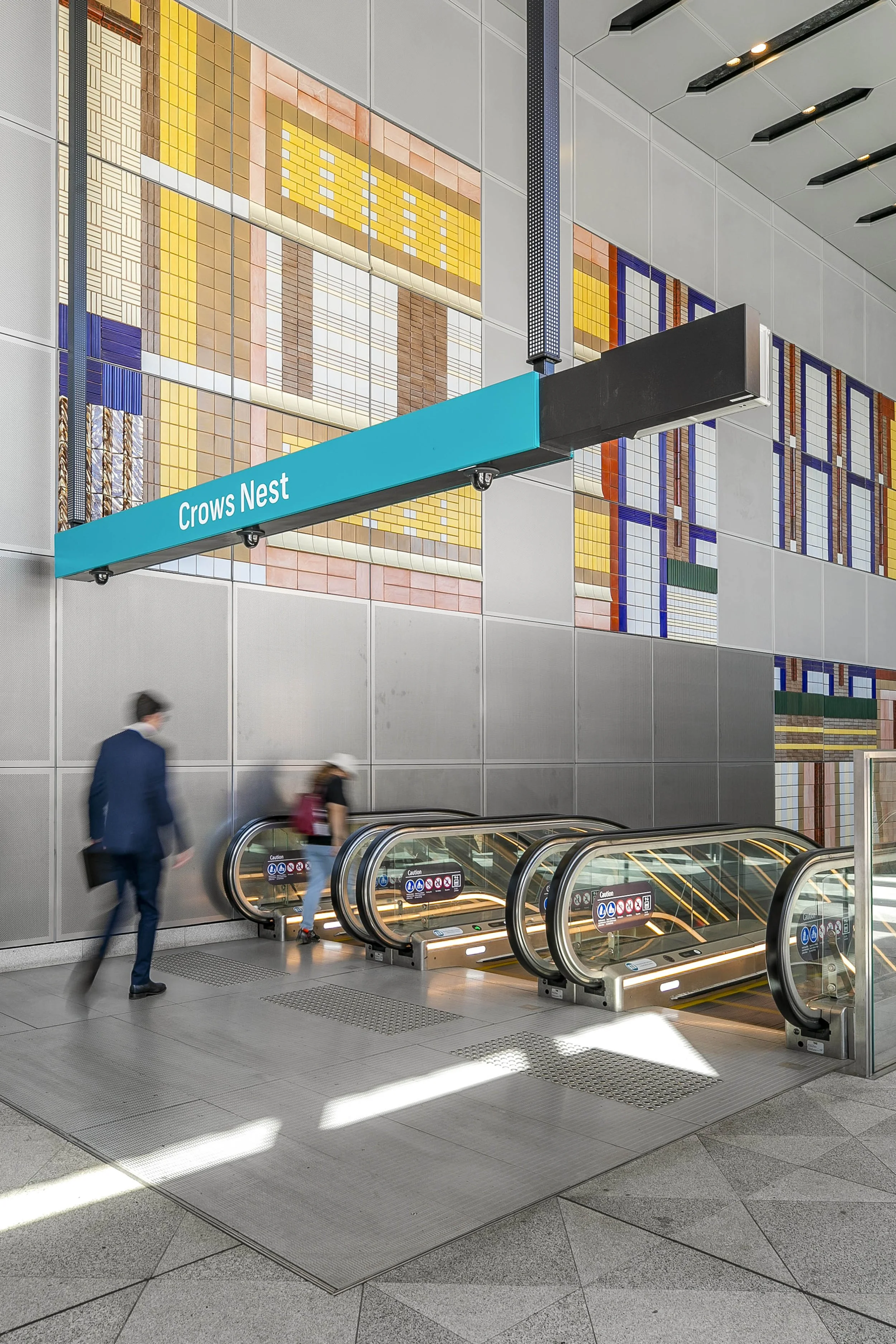 Crows Nest Metrol entrance with a blue sign reading 'Crows Nest,' escalators, and a large colourful mosaic wall.
