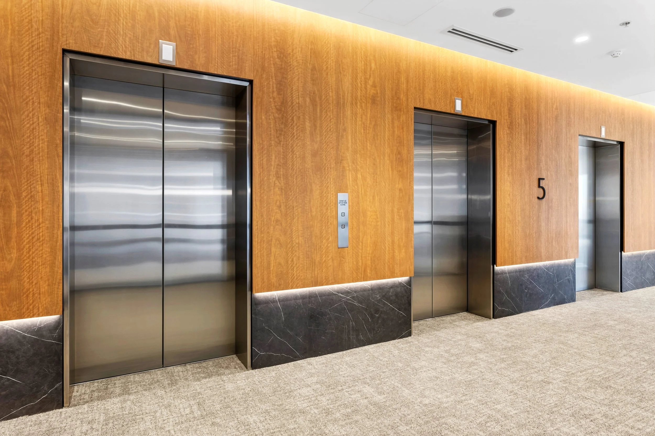 Three elevators with stainless steel doors in a modern hotel lobby, with wooden wall panels, a black marble baseboard, and a beige carpeted floor.