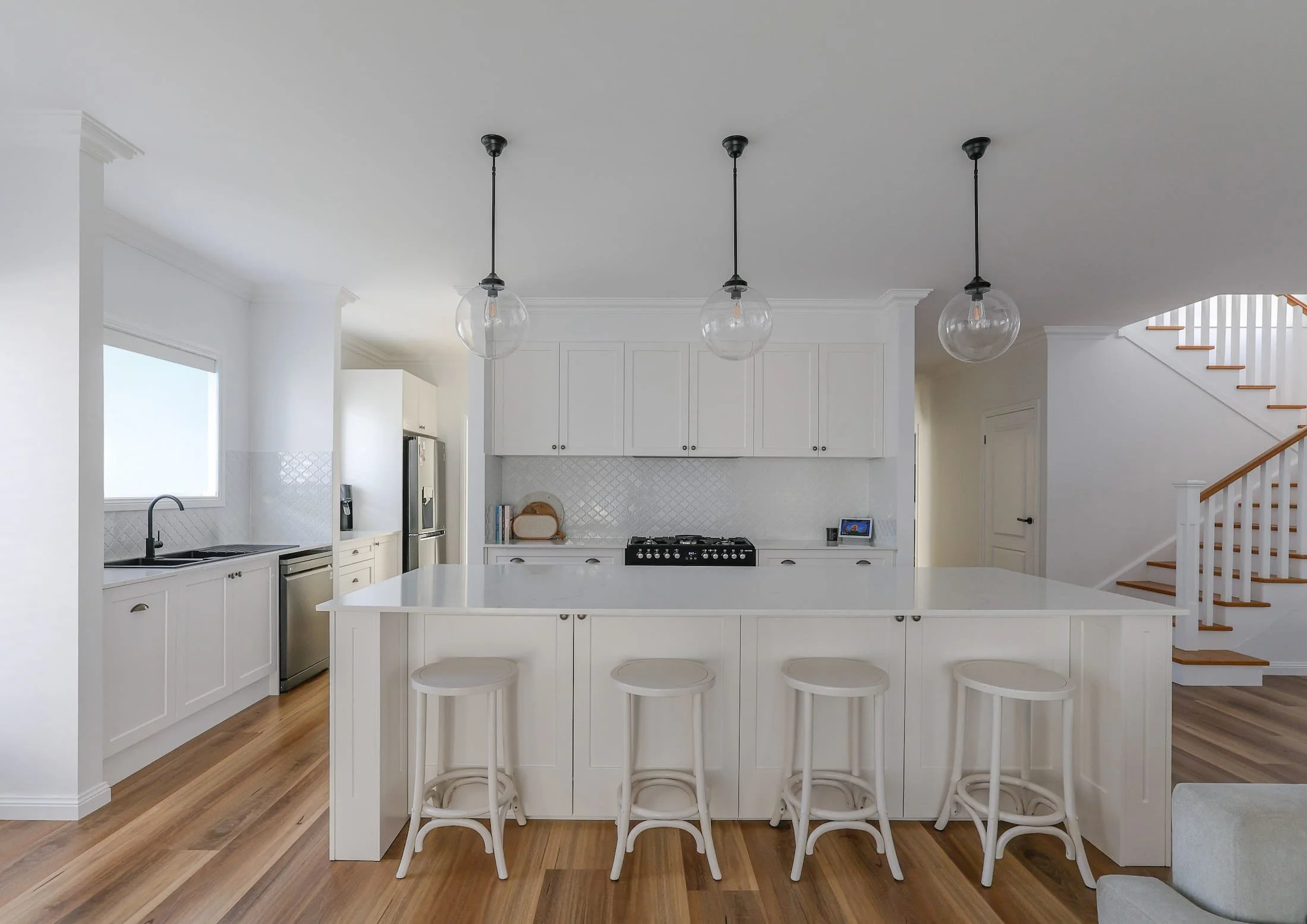 Modern white kitchen with a large island, three pendant lights, and a staircase to the right