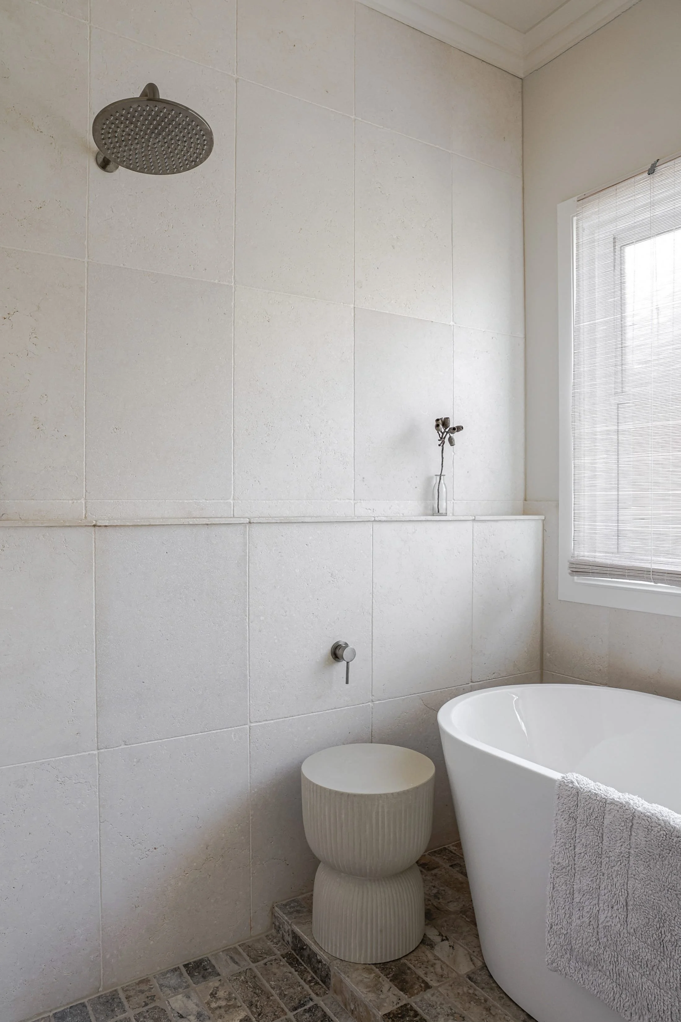 A modern bathroom featuring a shower with a rainfall showerhead, a large window with blinds, a white bathtub with a towel, and a textured stool next to it, with beige tiled walls and a patterned stone floor.