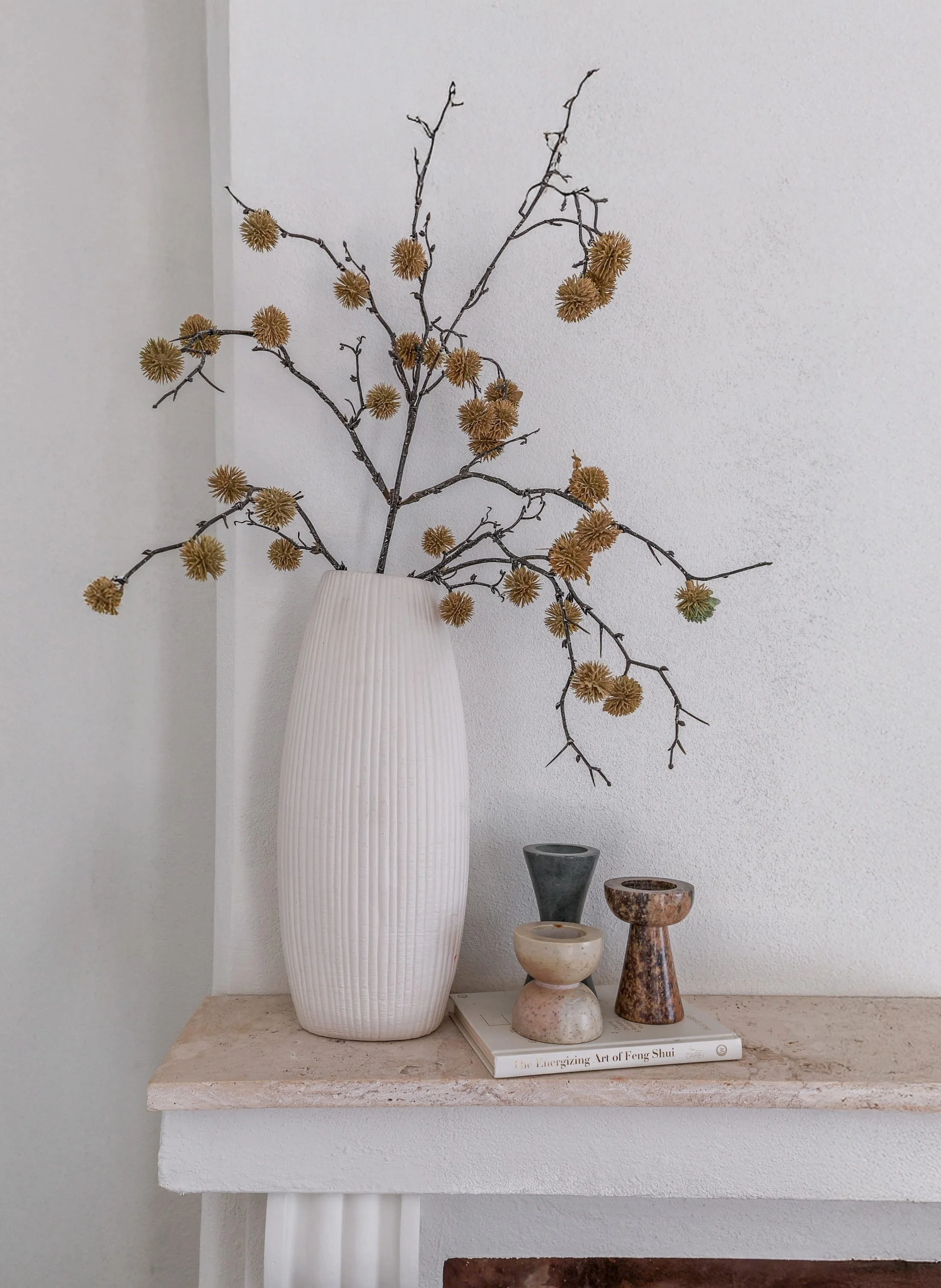 Decorative arrangement on a white mantel featuring a tall white vase with dried branches and seed pods, along with three small stone or ceramic vessels and a book titled "The Energizing Art of Feng Shui".