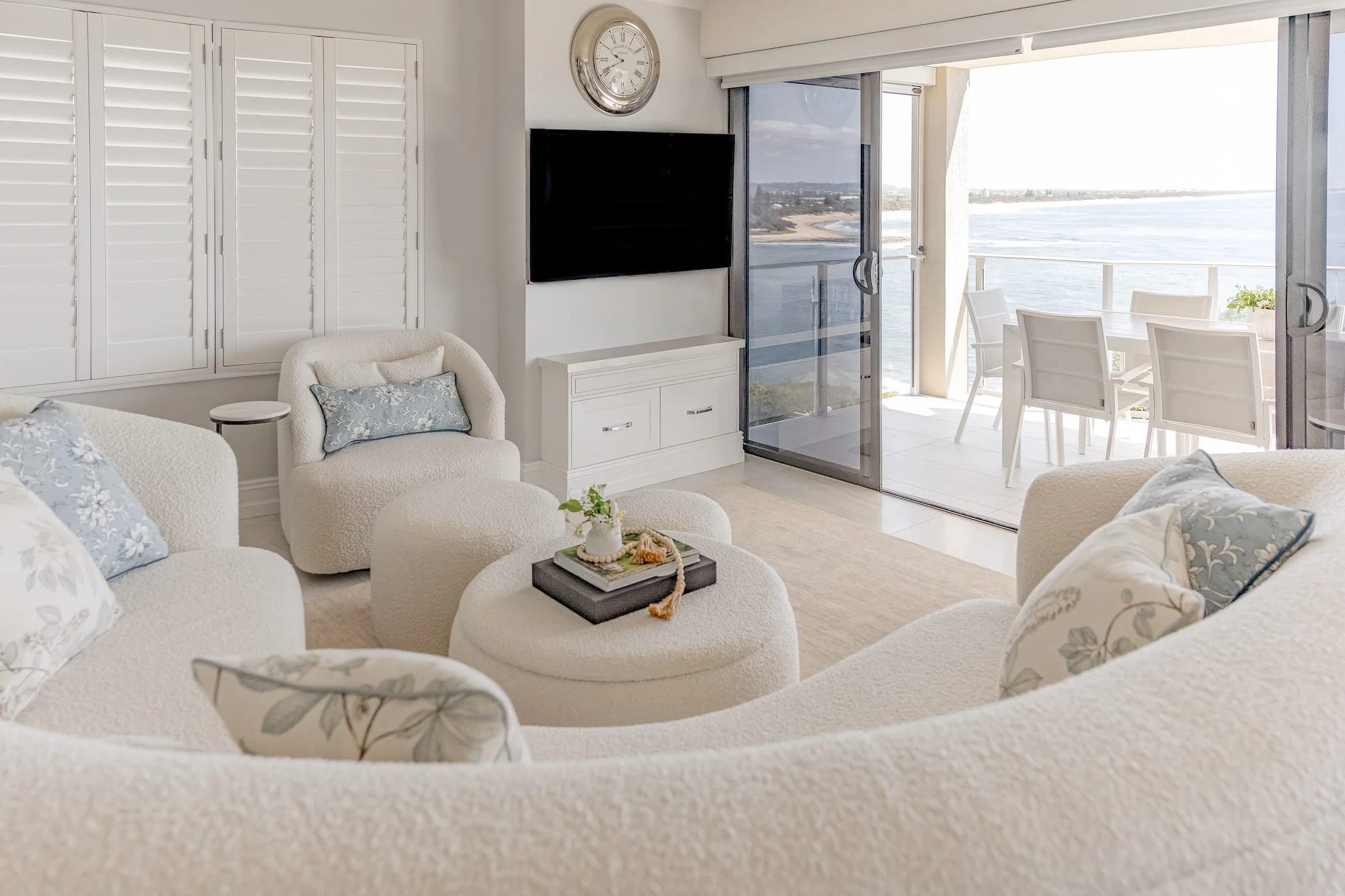 Living room with white upholstered furniture and pillows, a flat-screen TV, and a sliding glass door leading to a balcony overlooking the ocean.