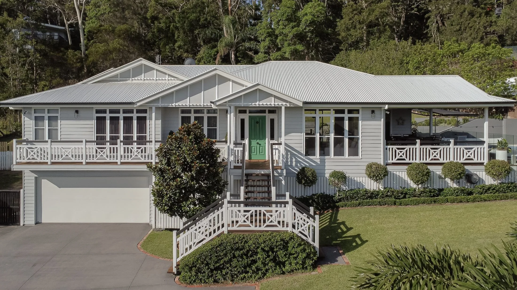 A modern white house with a metal roof, multiple decks, and a front staircase with railings, surrounded by a well-maintained lawn and landscaped shrubs, nestled in a green, wooded hillside.