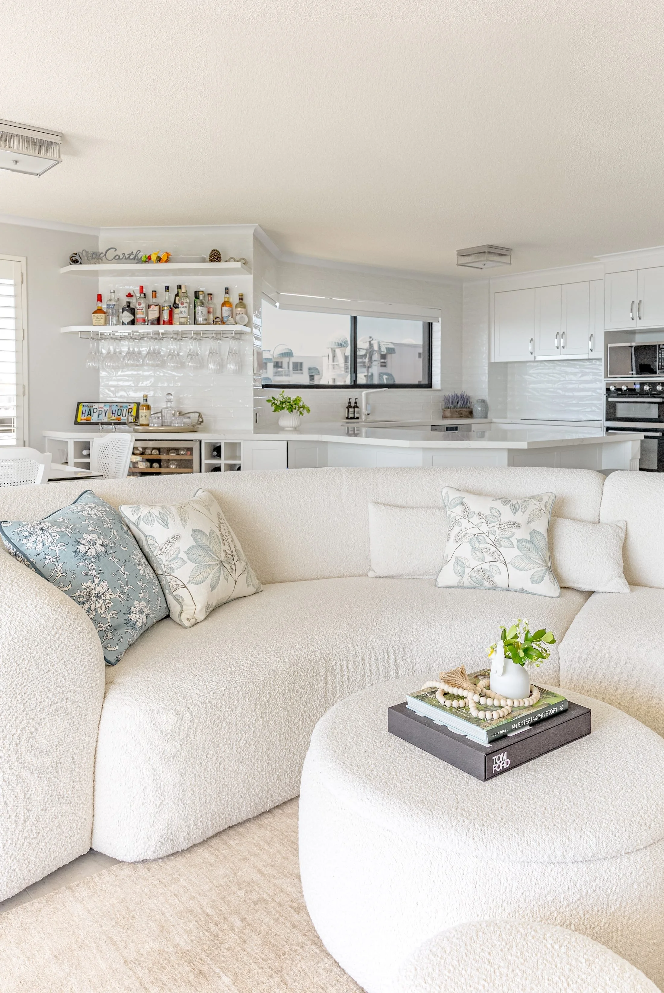A cozy living room with a cream-colored sectional sofa decorated with blue and floral pillows. On a matching round ottoman, there are magazines, a small vase with greenery, and decorative beads. In the background, an open kitchen features white cabinets, a window, and a bar area with liquor bottles and glassware.
