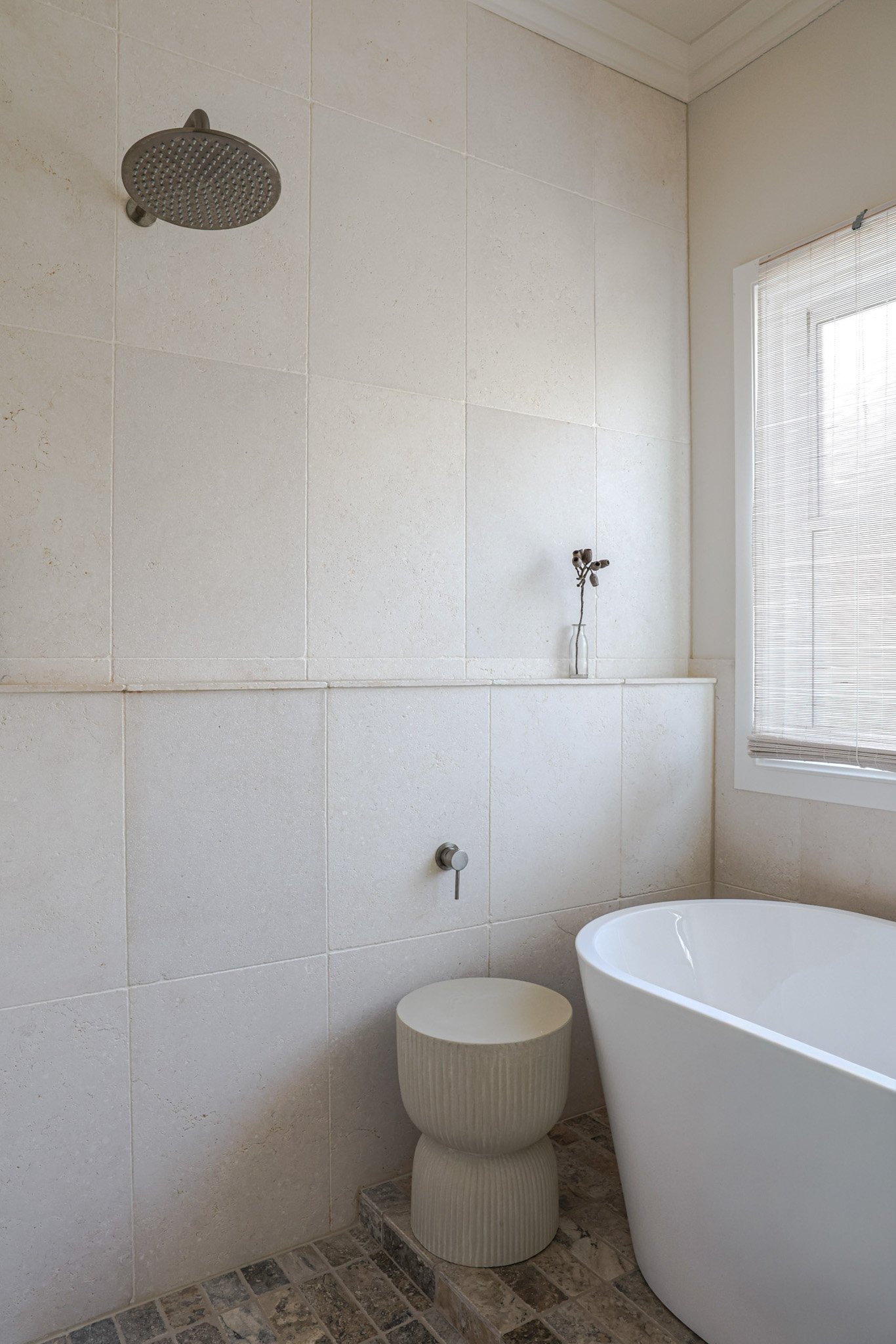 A modern bathroom featuring a shower with a rainfall showerhead, a large window with blinds, a white bathtub with a towel, and a textured stool next to it, with beige tiled walls and a patterned stone floor.