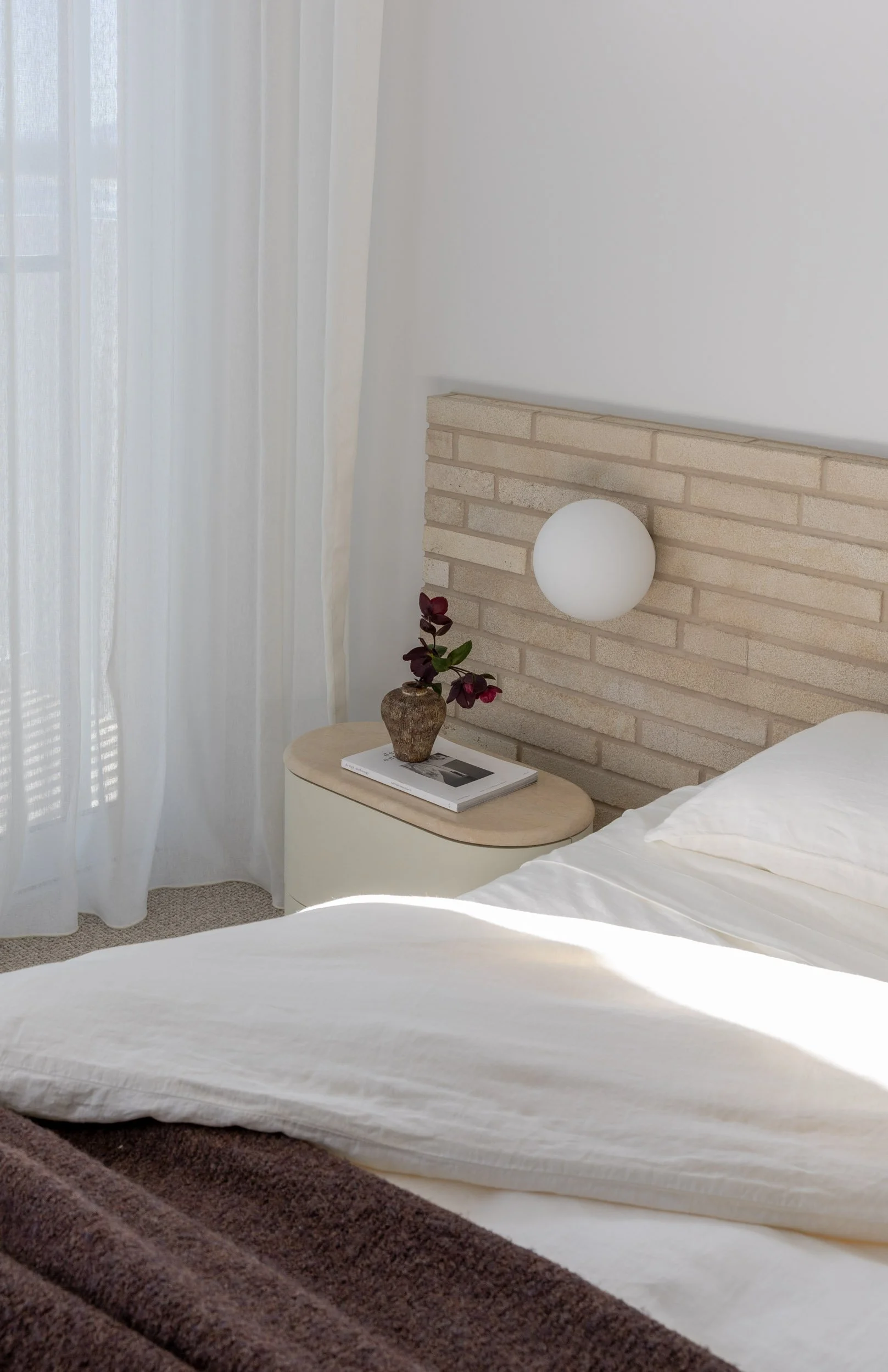 Bedroom with white brick feature, white walls, linen bedding, fresh maroon flowers, maroon throw, white linen sheers and round feature wall sconce
