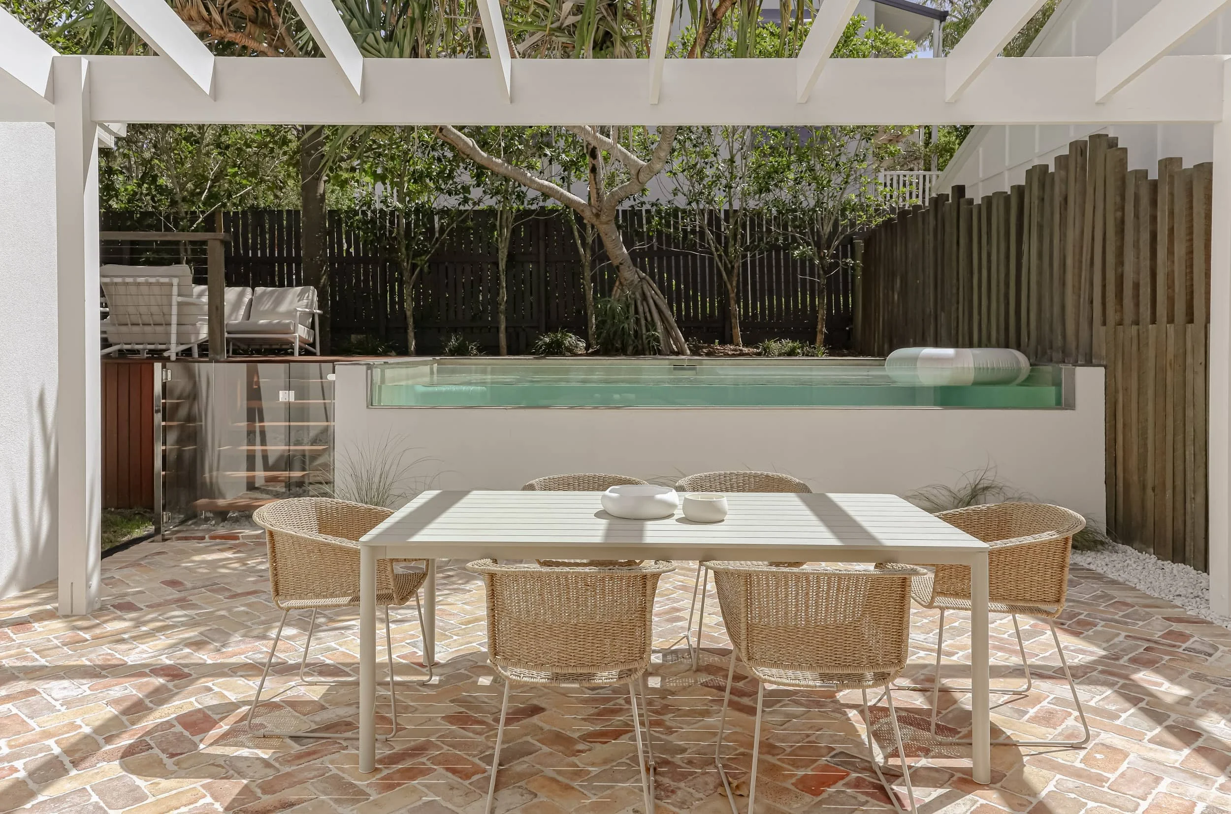 recycled red brick pavers, swimming pool under pandanus tree, dining table and wicker chairs, white open pergola, deck with seating area, striped sunny life pool ring, timber fence feature wall, glass pool fencing, grey and white outdoor setting