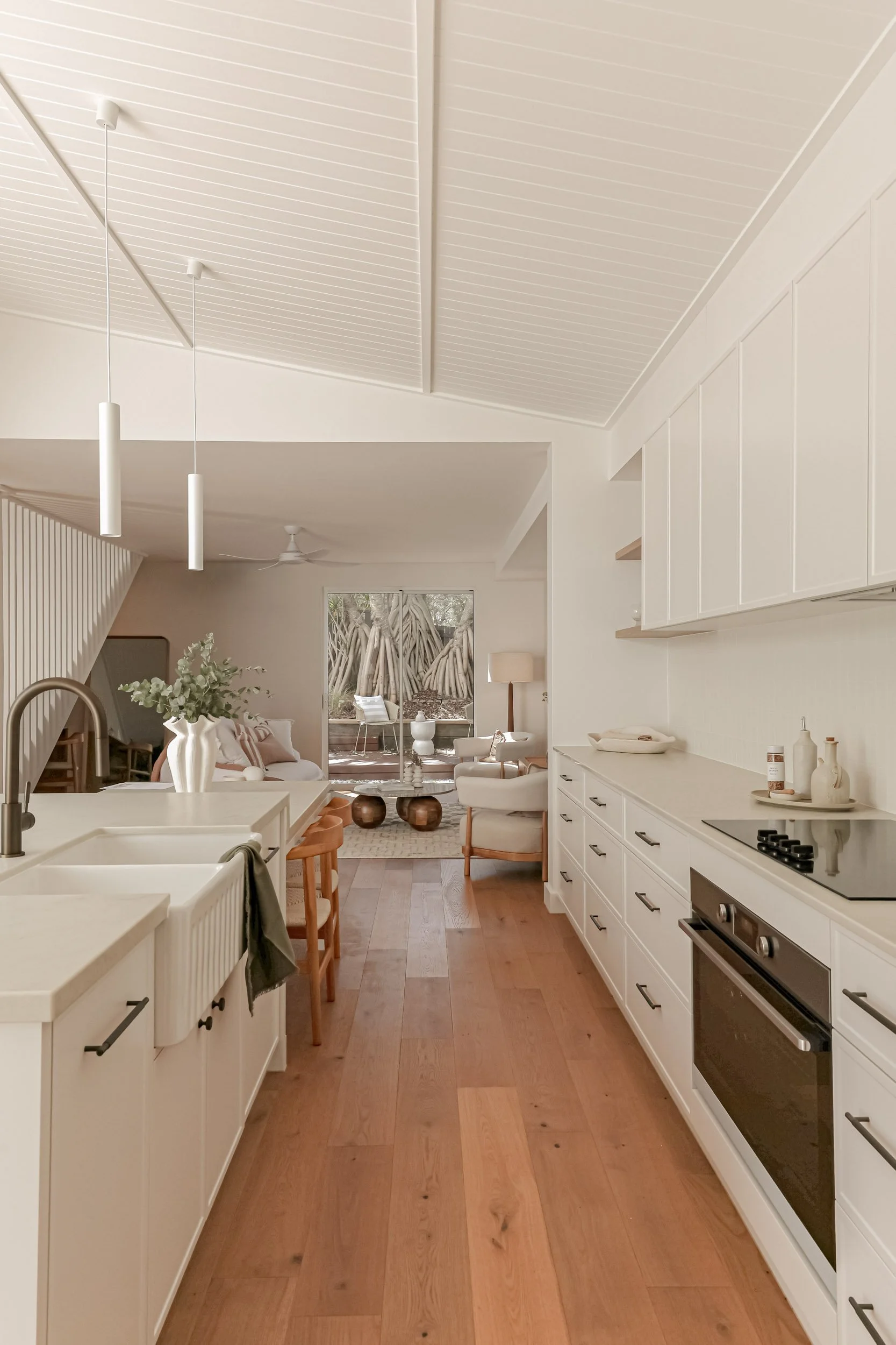 Modern open plan kitchen, dining and living, white slim shaker cabinetry, antique brass tap and handles, built in oven, white pendant lights, taj mahal stone bench, white vase with eucalyptus leaves, neutral styling, open timber shelves, occasional c