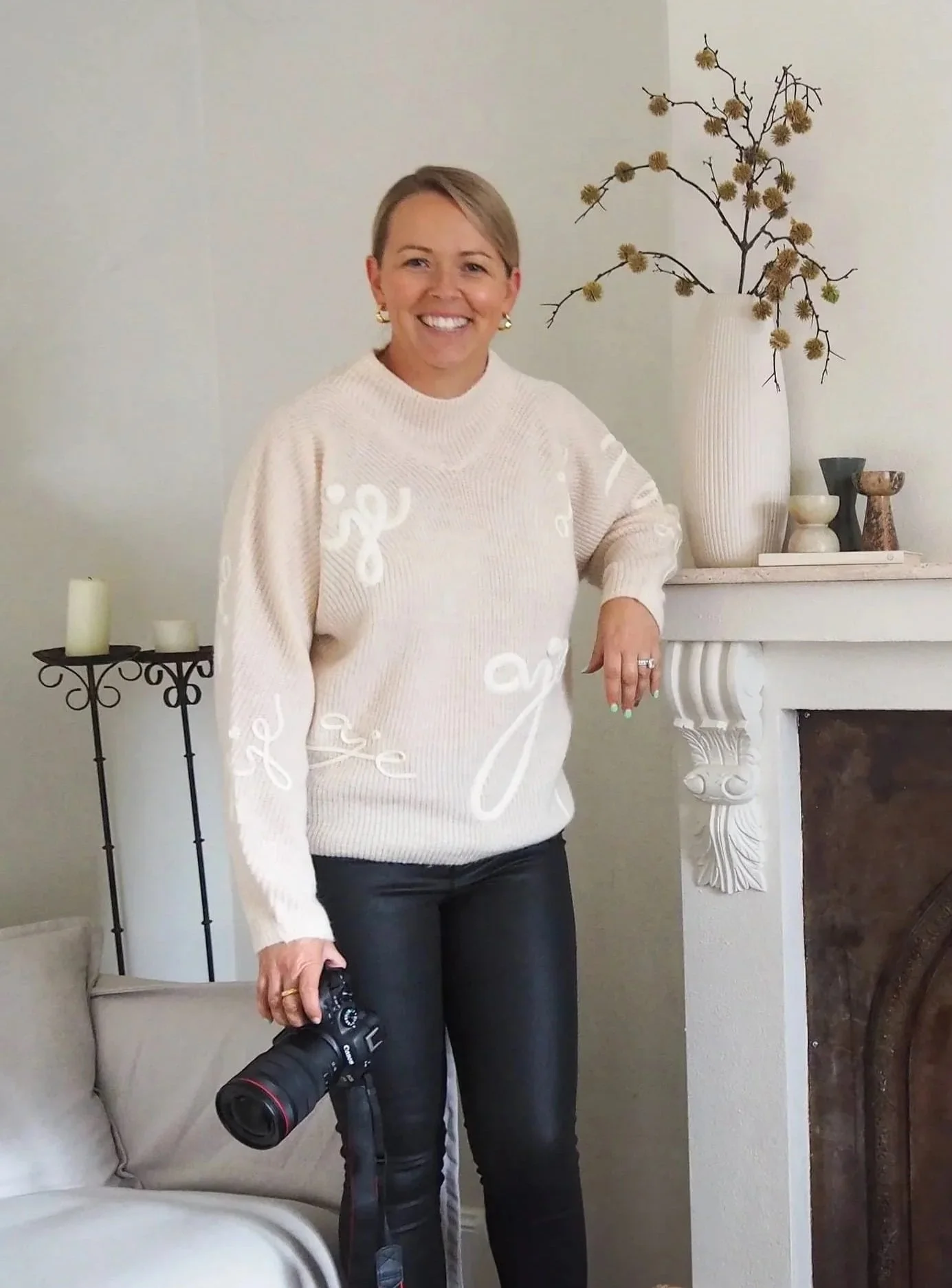 A woman smiling, holding a camera, standing next to a fireplace with a large vase and dried branches on the mantelpiece, in a well-lit room with white walls.
