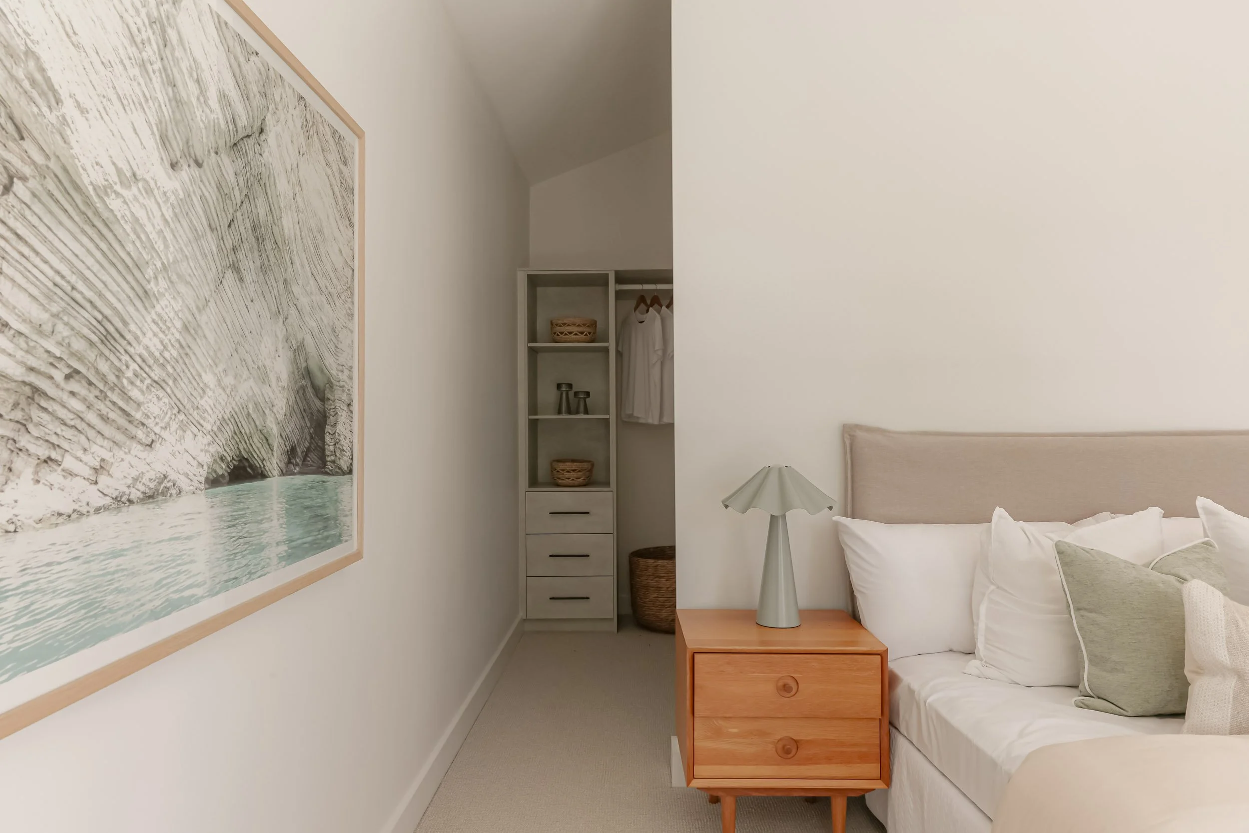 Bedroom with walk in robe, sage green pillows and lamp, timber bedside table, light grey wardrobe joinery, light carpet, white bed linen, coastal artwork 