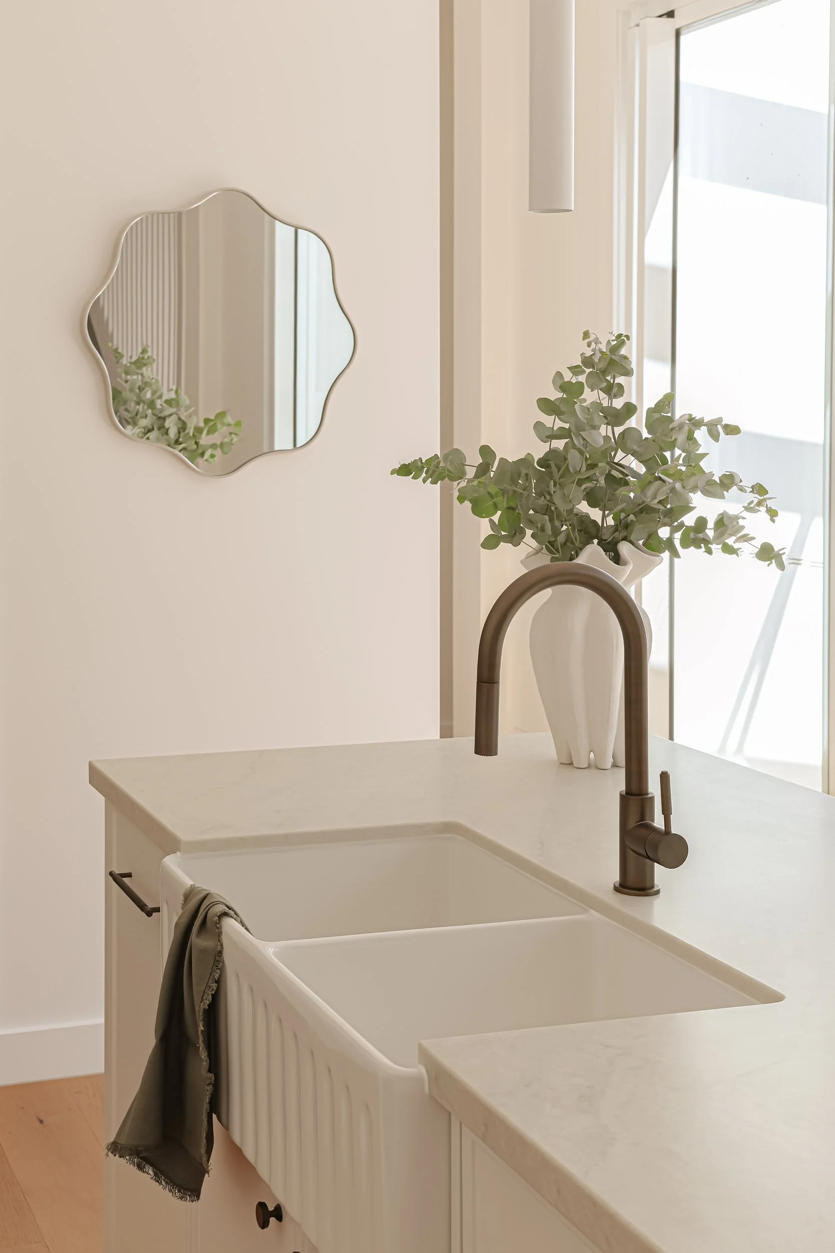 Taj mahal stone bench top, antique brass tap, white vase with eucalyptus, green linen hand towel, mirror, white pendant light, light timber floor, white farmhouse sink