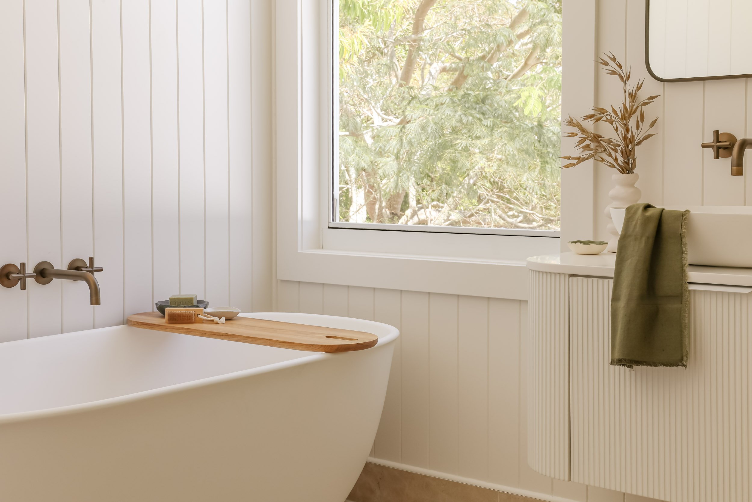 Antique brass mirrors, antique brass tapware, white fluted vanity, white stone bench top, green linen hand towel, white VJ wall panelling, white cabinet handles, white bath, timber bath board with soap and timber scrubbing brush, window with greenery