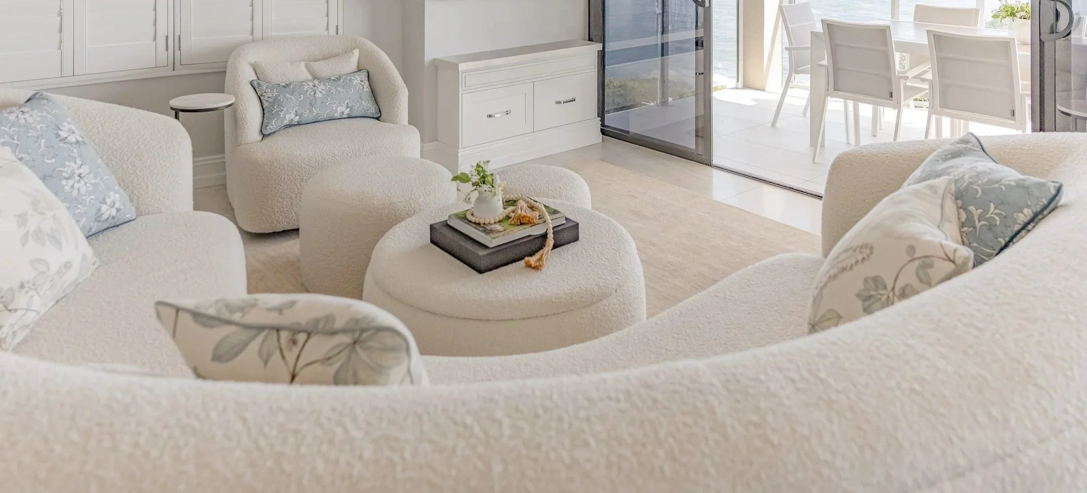 Living room with white upholstered sofa and chairs, floral throw pillows, and a black tray with decorative items on an ottoman, with a view of an outdoor dining area through a glass sliding door.
