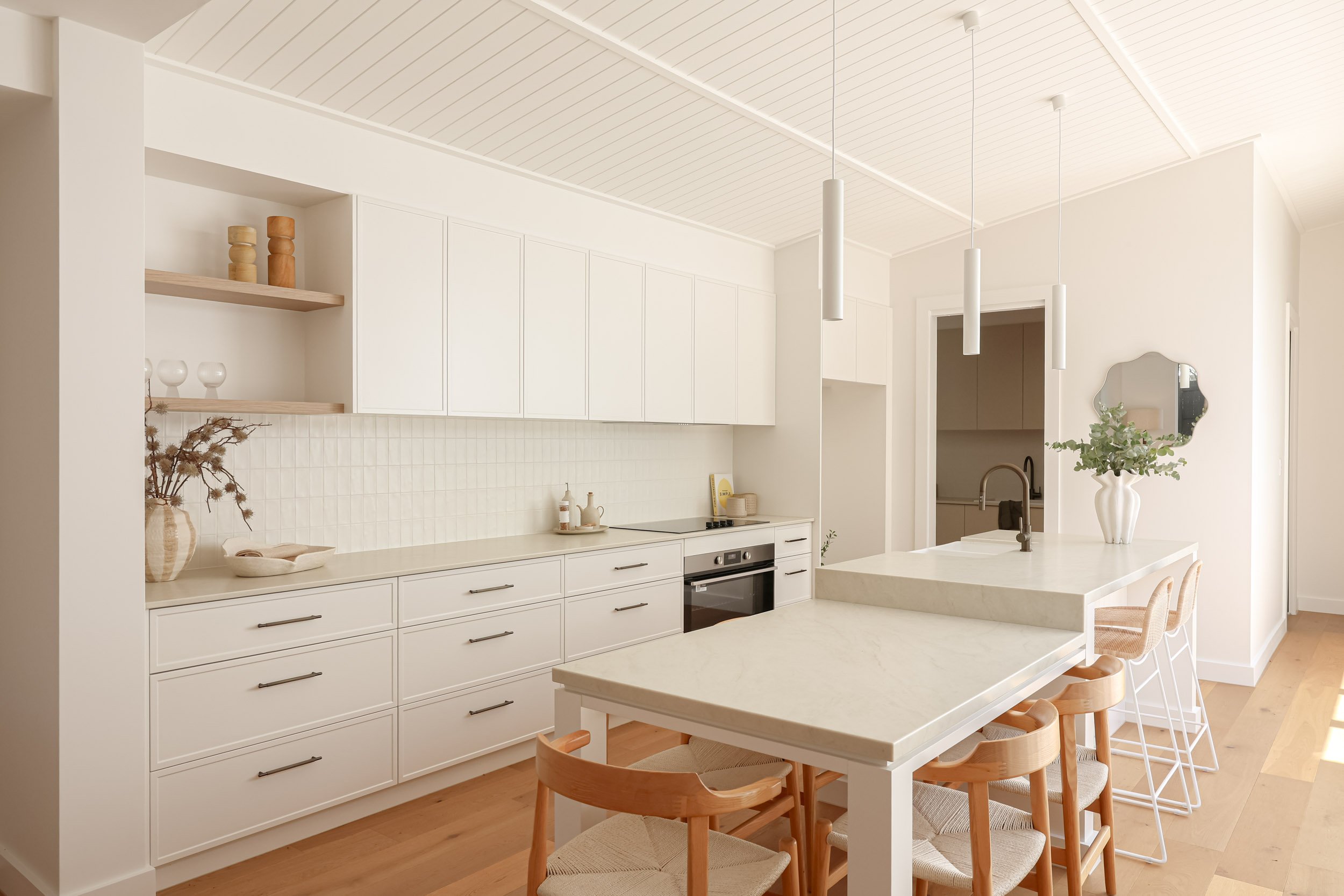 White kitchen, shaker cabinets, taj mahal stone, antique brass tapware, light timber shelves, mirror, white pendant lights, timber dining chairs, greenery in vase, vj board ceiling