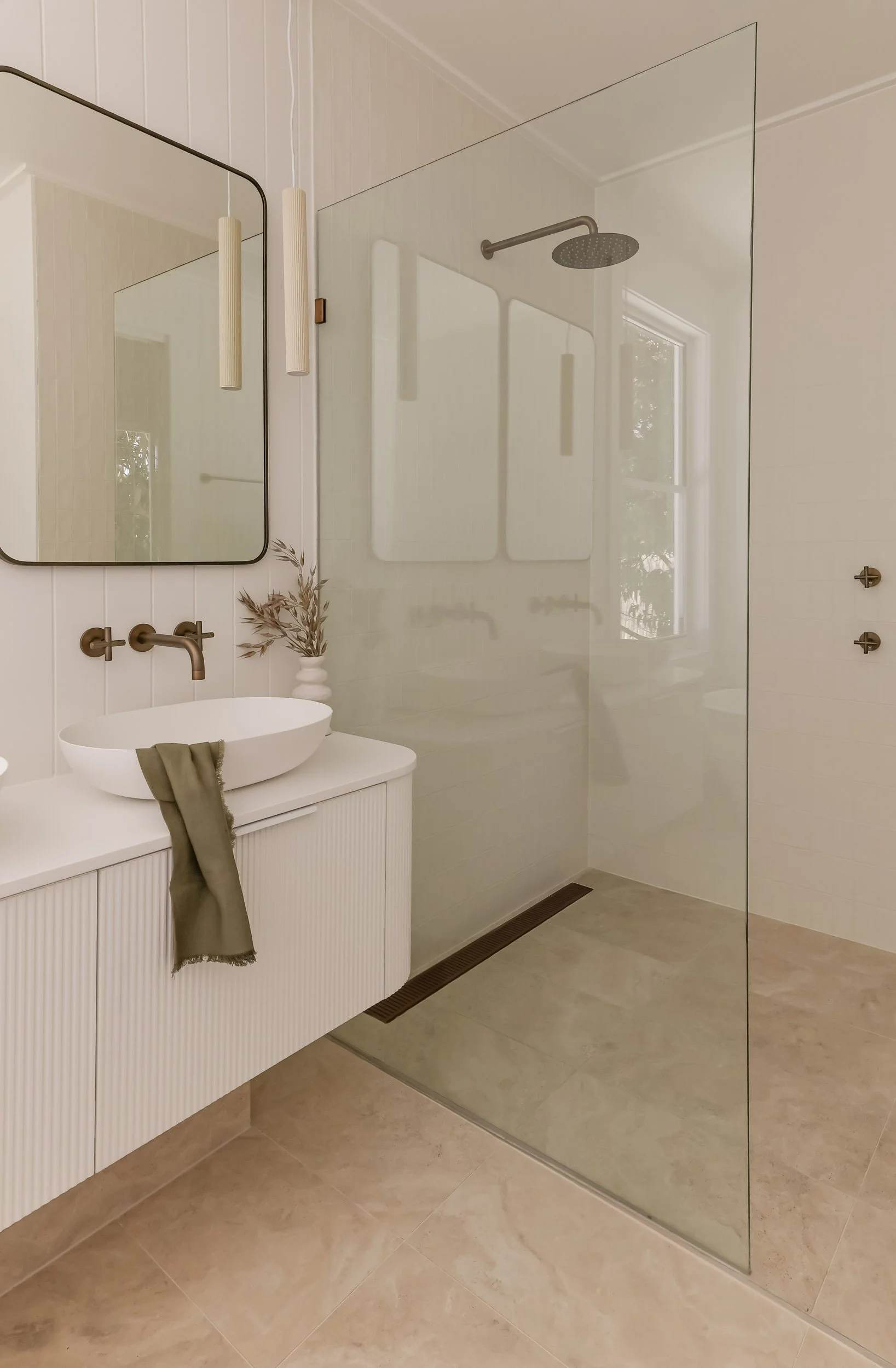 Antique brass mirrors, antique brass tapware, white fluted vanity, white stone bench top, green linen hand towel, white VJ wall panelling, white cabinet handles, large open shower, beige tiles, small white vase with greenery