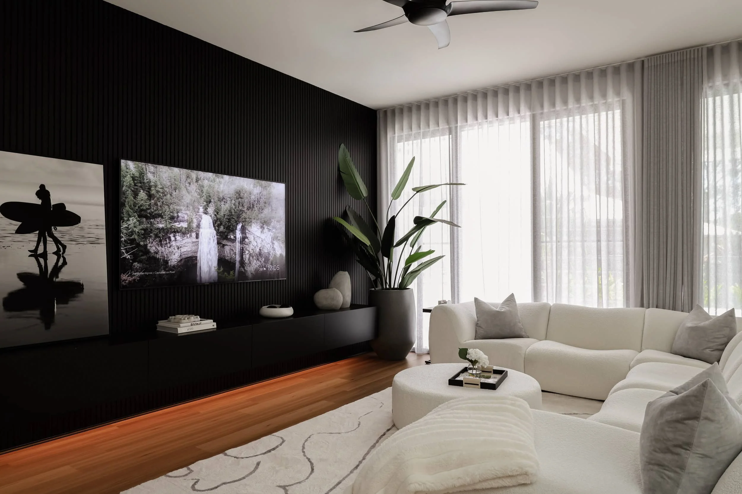 Modern living room with black timber feature wall, light green linen sheers, white boucle couch, white and grey rug, grey cushions, bird of paradise plant