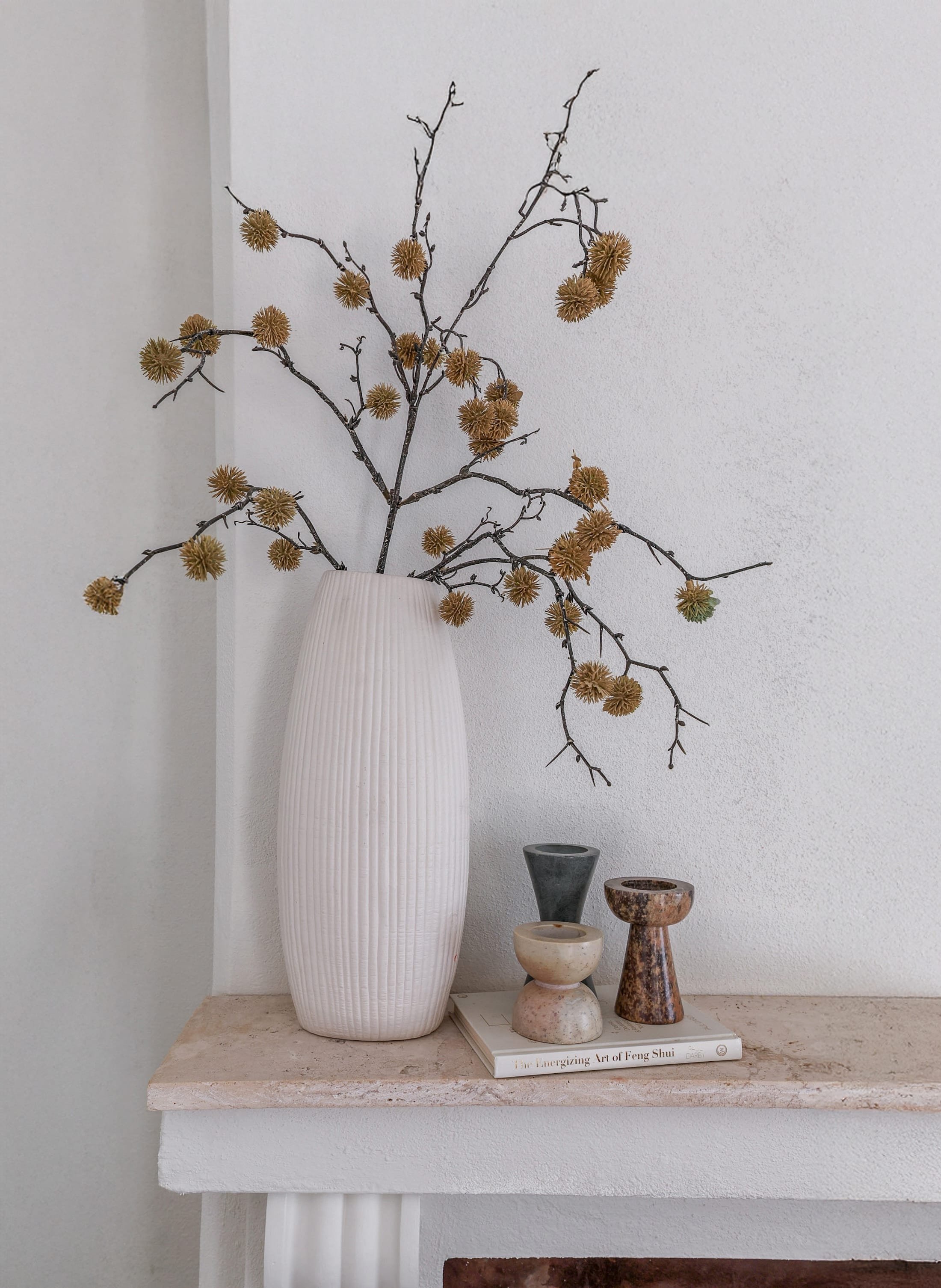 A white ribbed vase with dried branches and seed pods inside, placed on a beige marble fireplace mantel. Next to the vase are three small decorative pottery pieces and a book titled 'The Energizing Art of Feng Shui'.