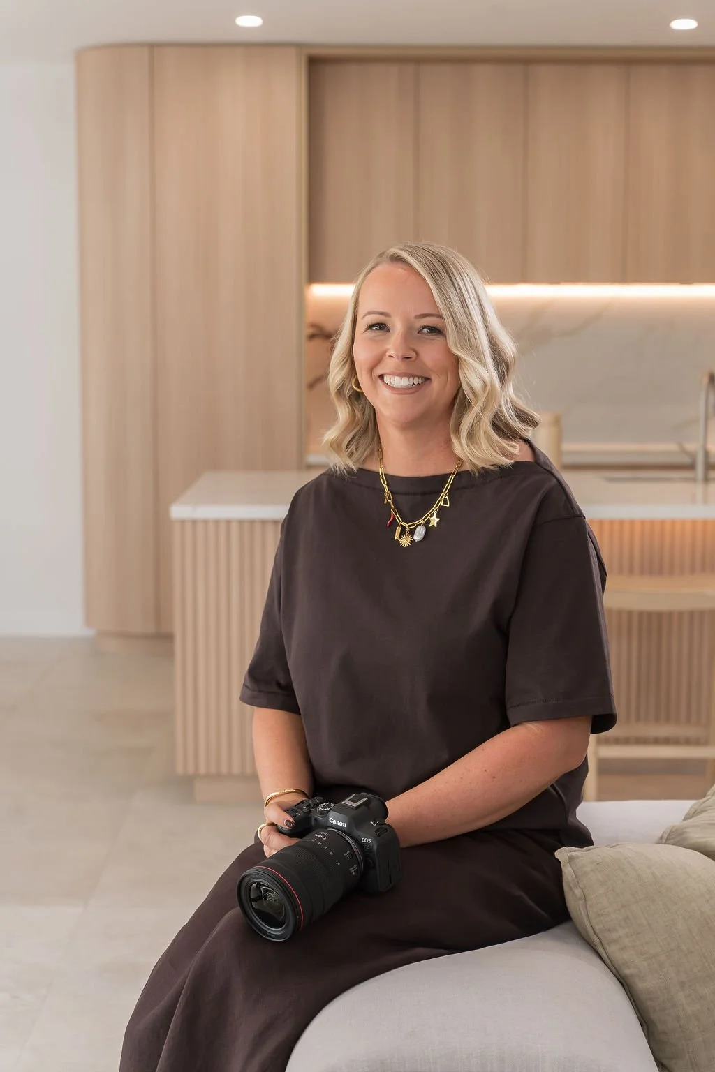 A woman smiling while holding a camera, sitting on a light grey sofa in a living room with a kitchen behind her, decorated with a timber stool, stone bench top, tall vase with branches on the benchtop