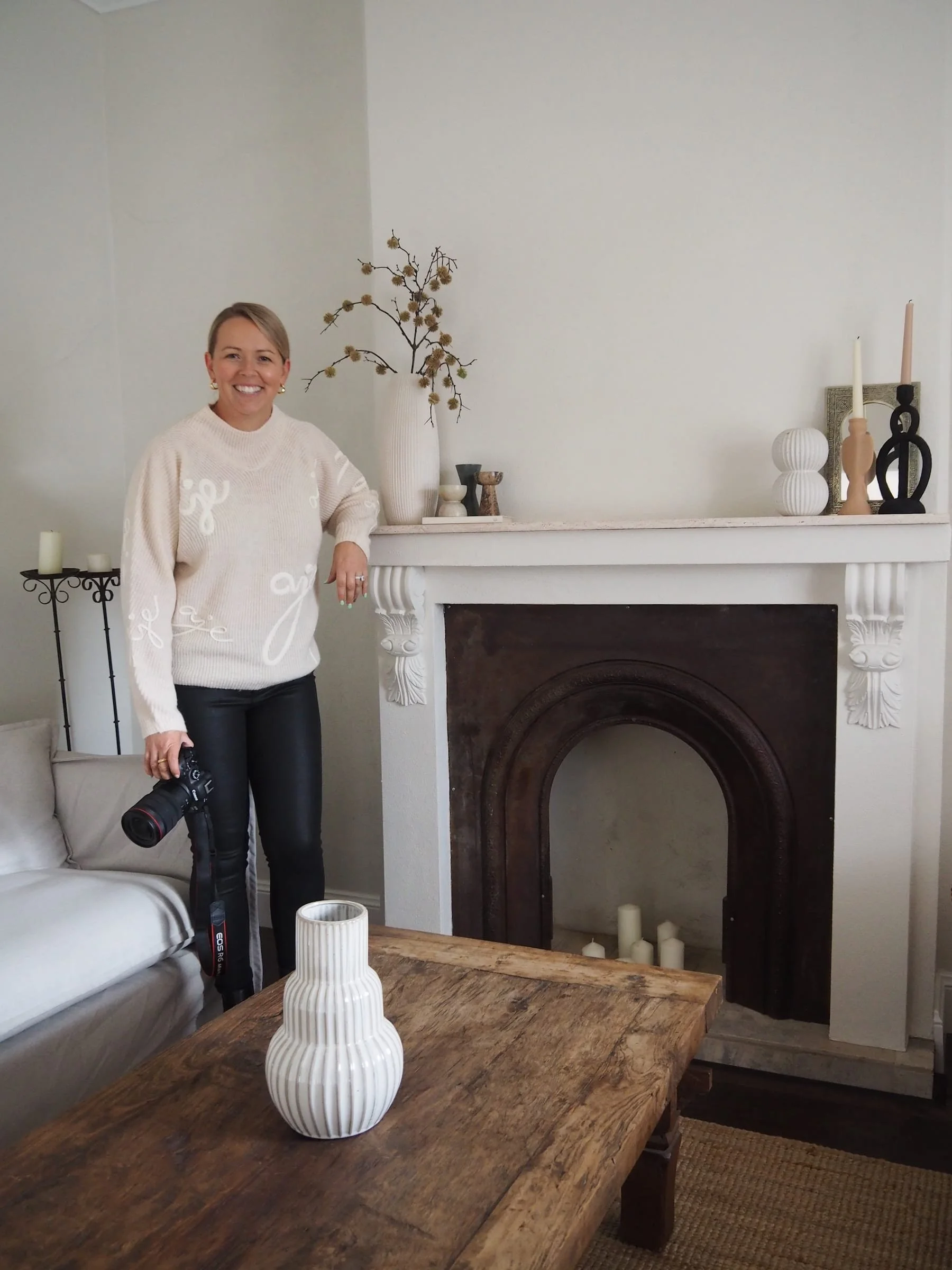 A woman smiling while holding a camera, standing next to a beige sofa in a living room with a fireplace, decorated with vases, candles, and a tall vase with branches on the mantelpiece.