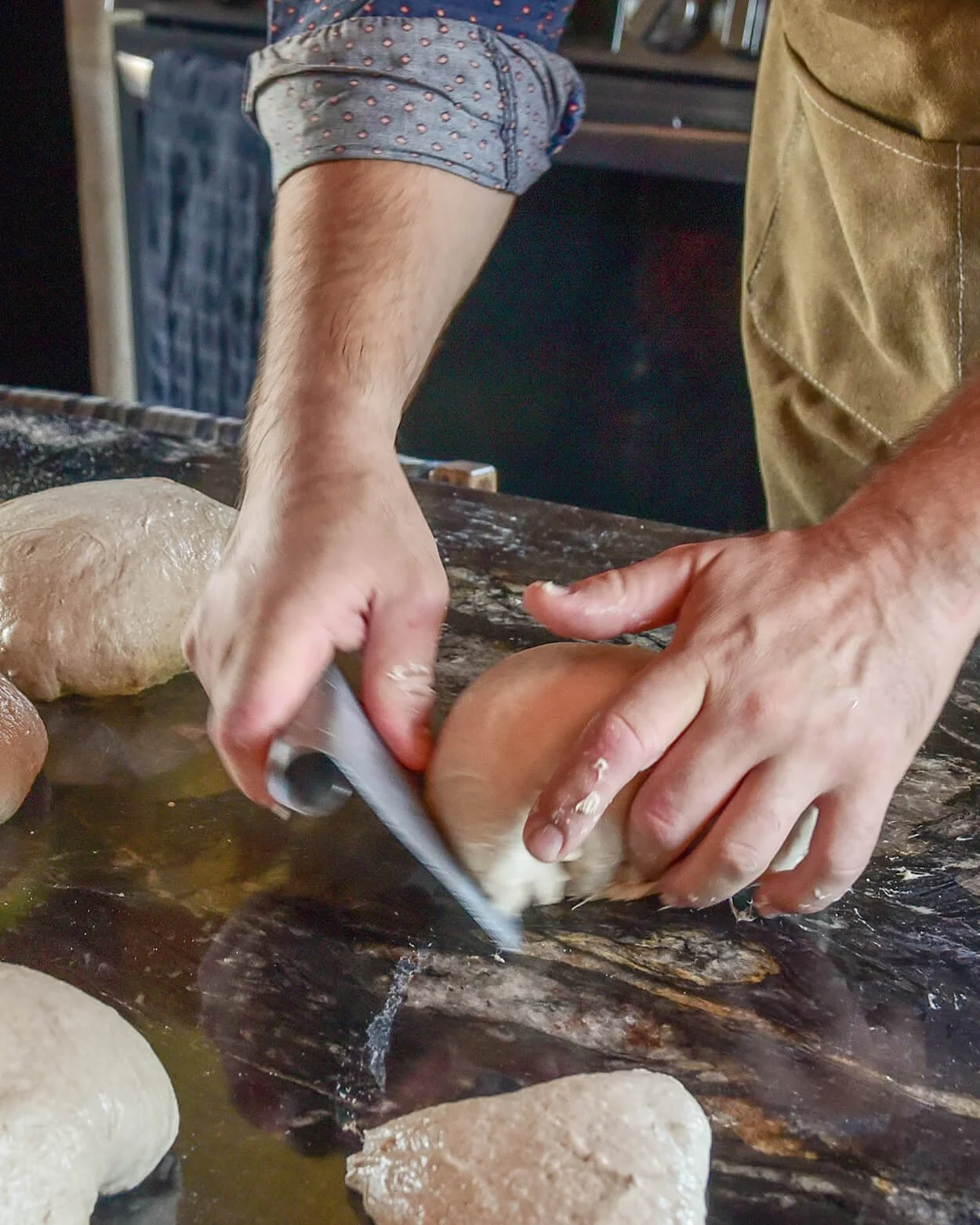 Person slicing dough with a pizza cutter on a wooden surface, with multiple dough balls around.