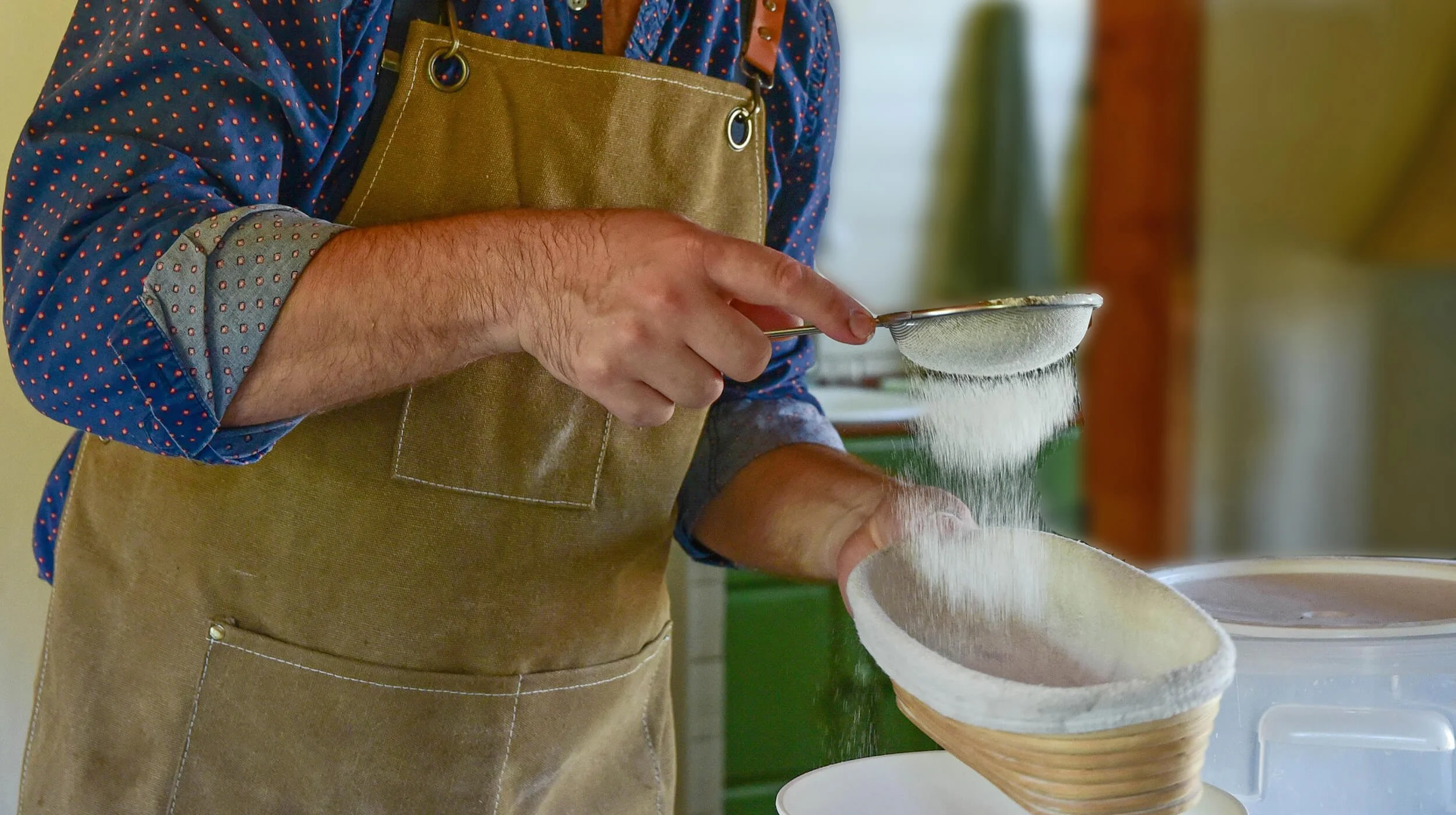 A person sifting flour through a metal sieve into a bowl, wearing a brown apron and a blue shirt with red dots.