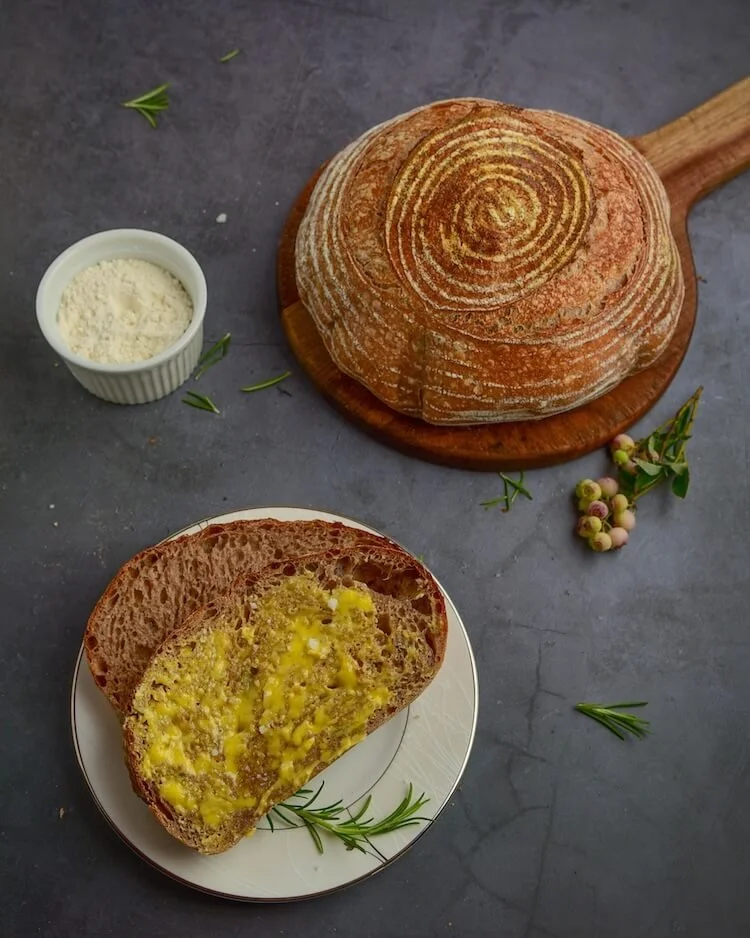 A loaf of bread on a wooden cutting board, a slice of bread with butter and herbs on a plate, a small bowl of grated cheese, and sprigs of herbs and small berries on a dark surface.
