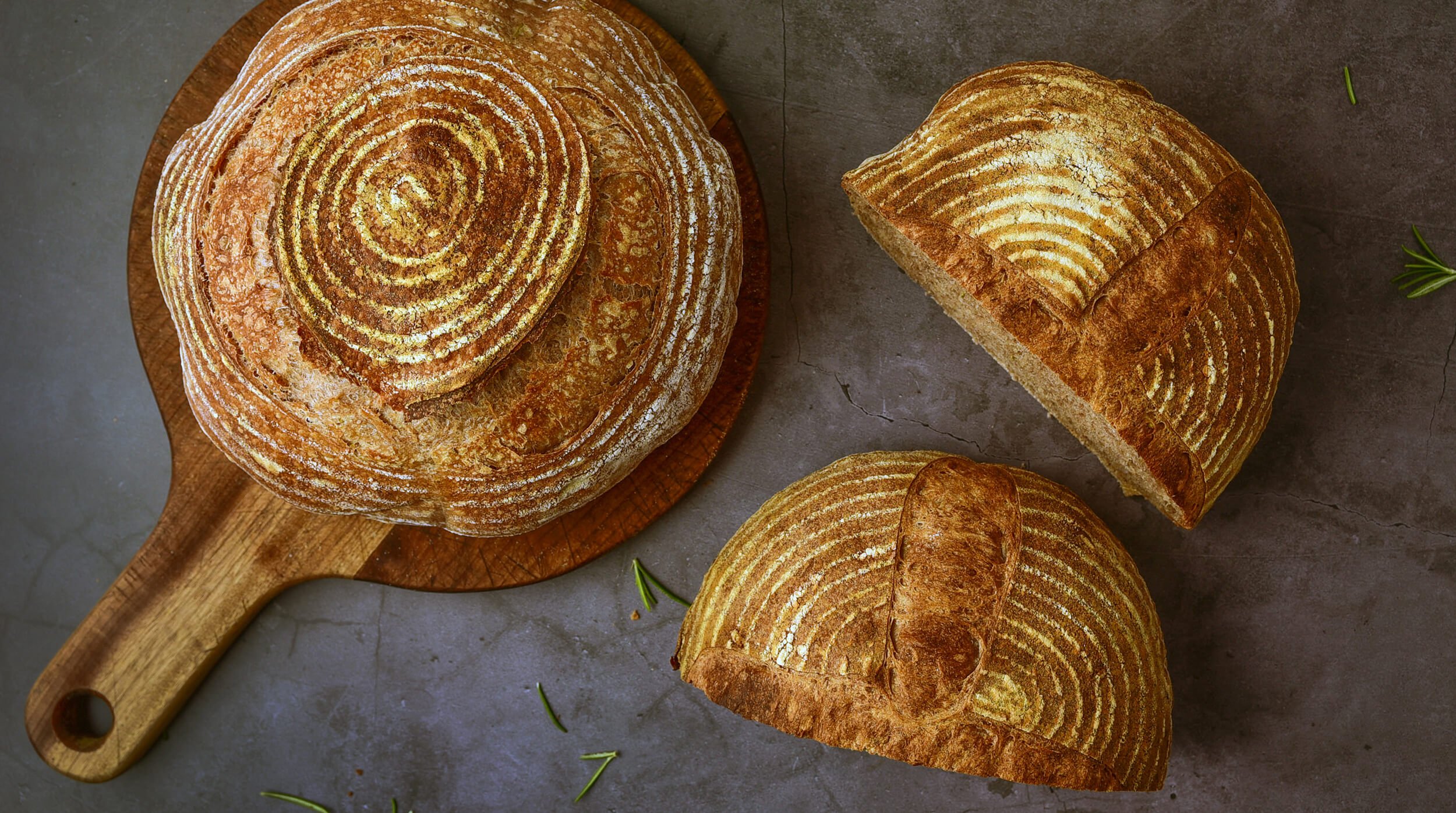 Round loaf of sourdough bread with concentric rings, placed on a wooden cutting board; two slices cut from the loaf and placed on a gray surface, with small green herbs scattered around.