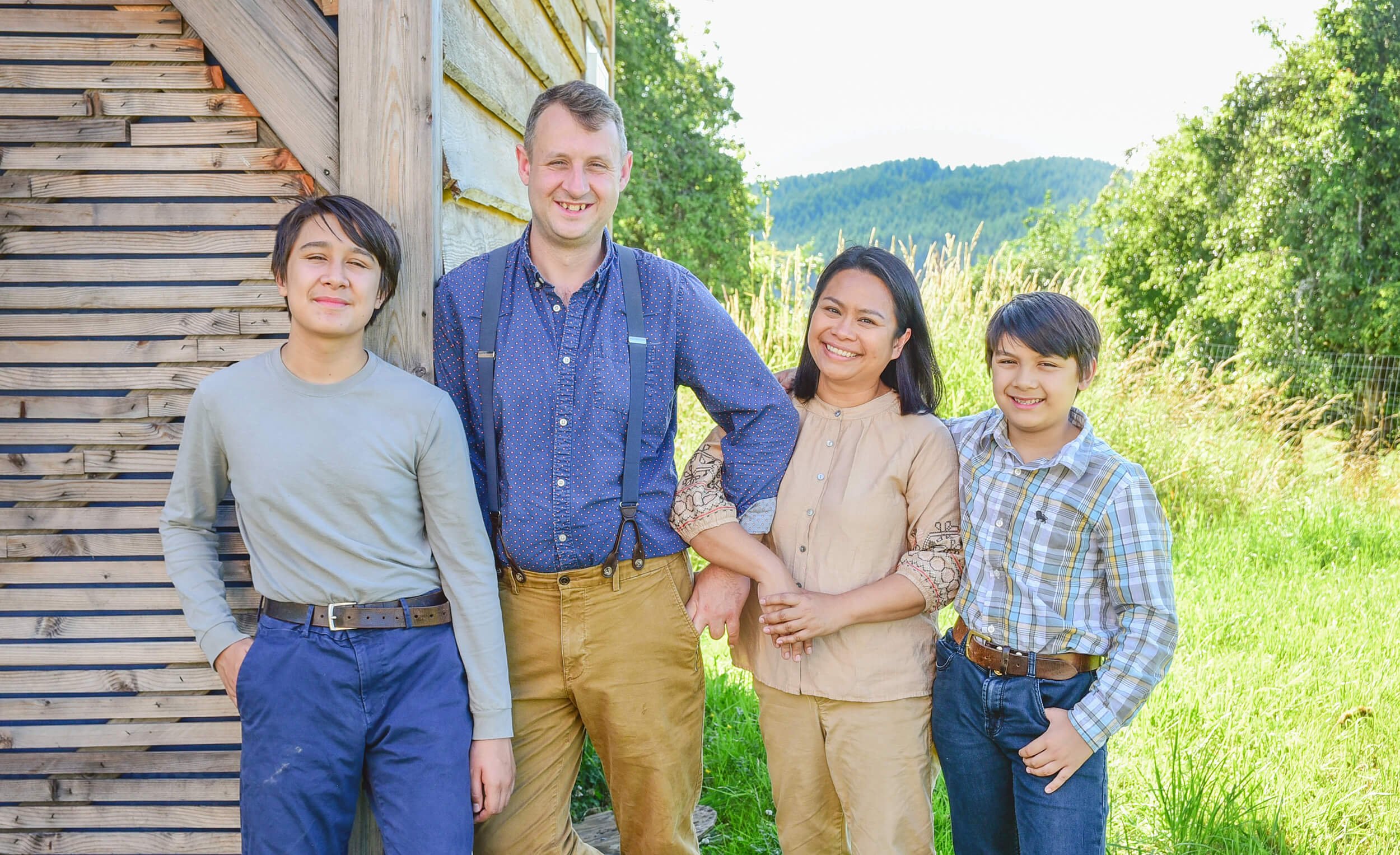 A diverse group of five people, a mix of adults and children, standing outdoors near a wooden structure with green trees and a grassy field in the background. They are smiling and appear to be enjoying a sunny day.