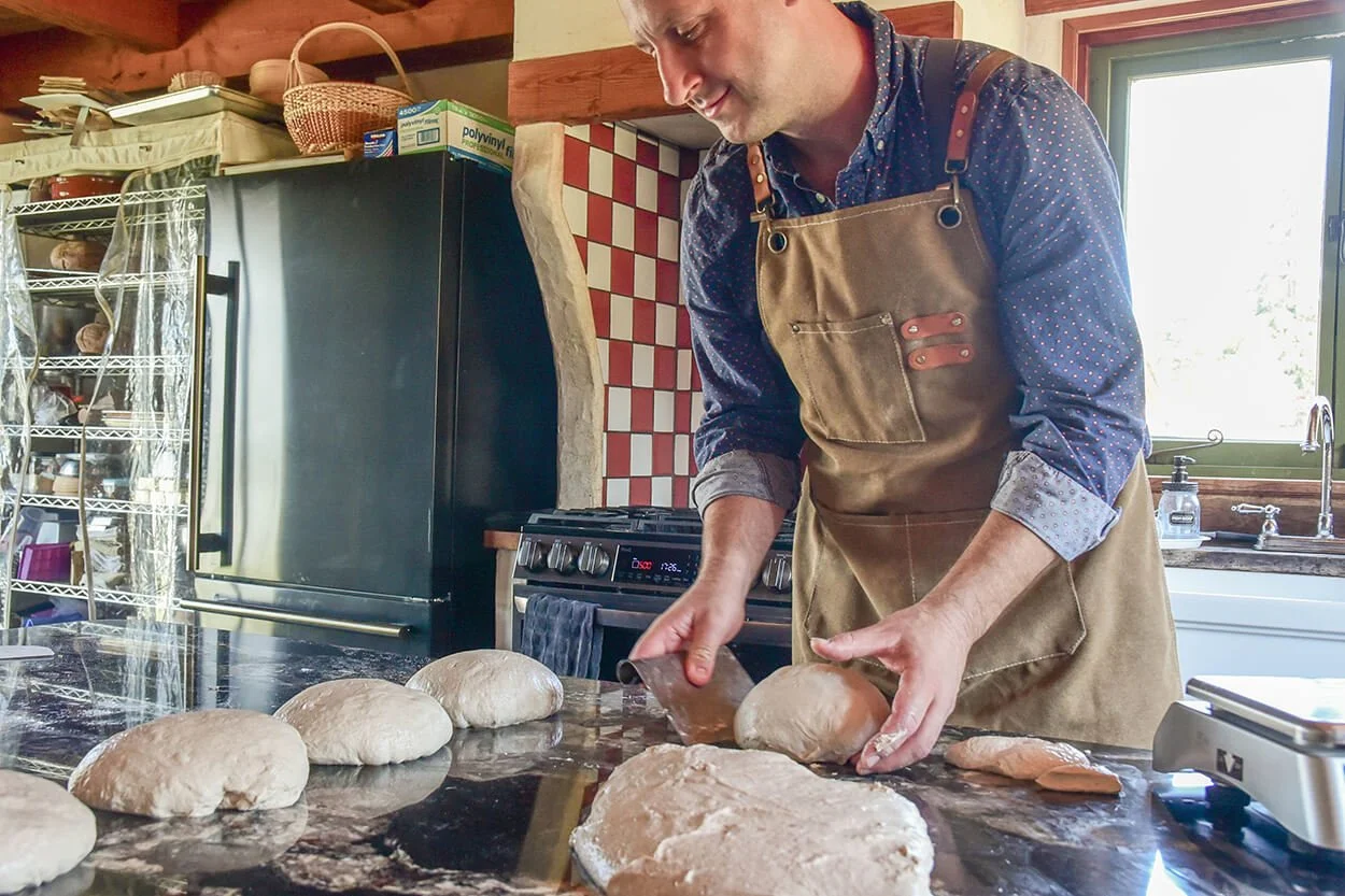 A man in a brown apron and blue shirt preparing dough on a kitchen countertop.