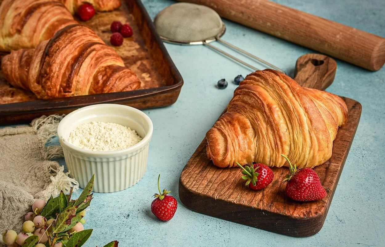 A croissant on a wooden serving board with strawberries beside it, a baking tray with more croissants and berries, a bowl of flour, a tea infuser, a rolling pin, and some berries and foliage decorate the light blue surface.