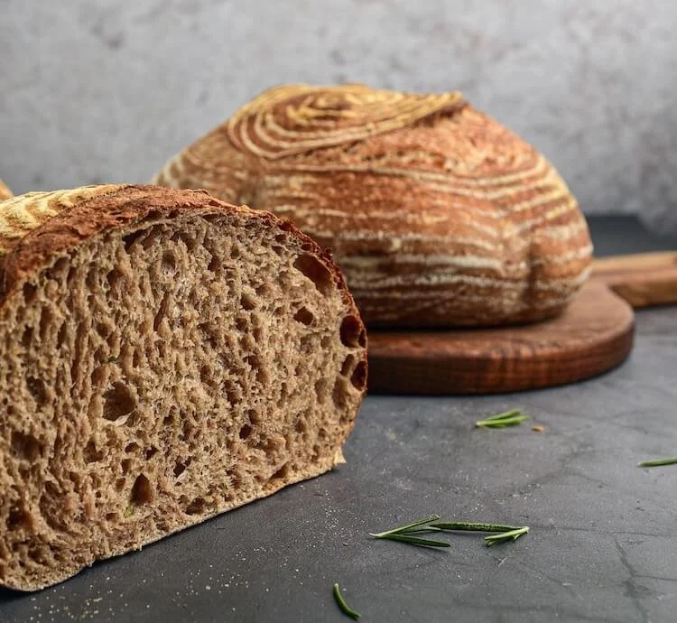 A loaf of bread with a slice cut, showing a dense and crumbly interior, placed in front of the whole loaf on a dark surface with some rosemary sprigs nearby.