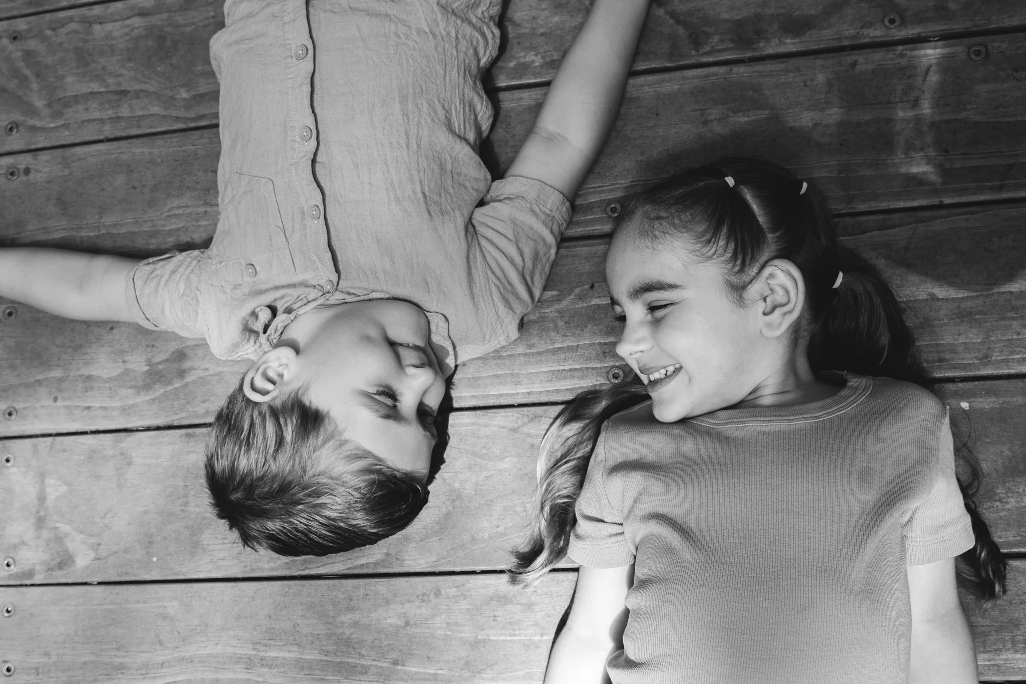 A boy and a girl lying on a wooden floor, facing each other, smiling and looking happy.