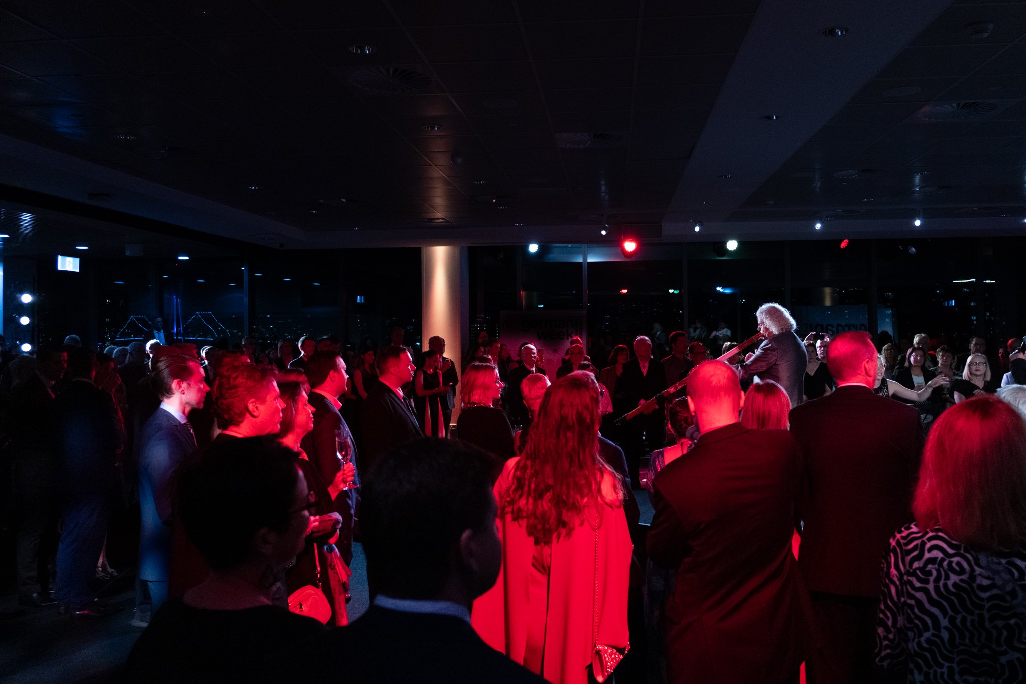 A live musical performance at an indoor event with a musician playing guitar on a stage to a large seated audience in formal attire, illuminated by red and blue lights.