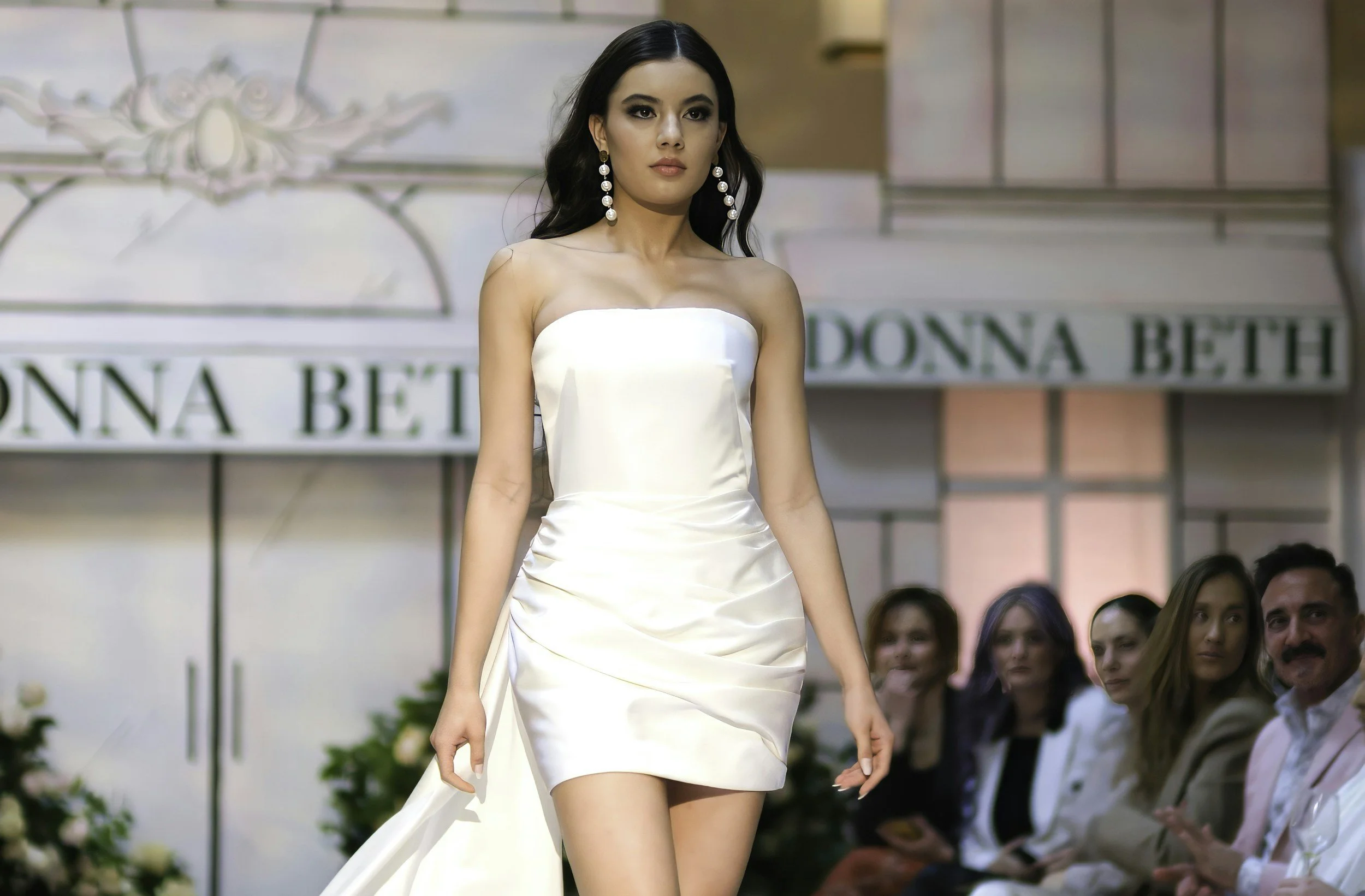 A woman walking on a runway during a fashion show wearing a strapless white dress and large pearl earrings, with an audience watching attentively.