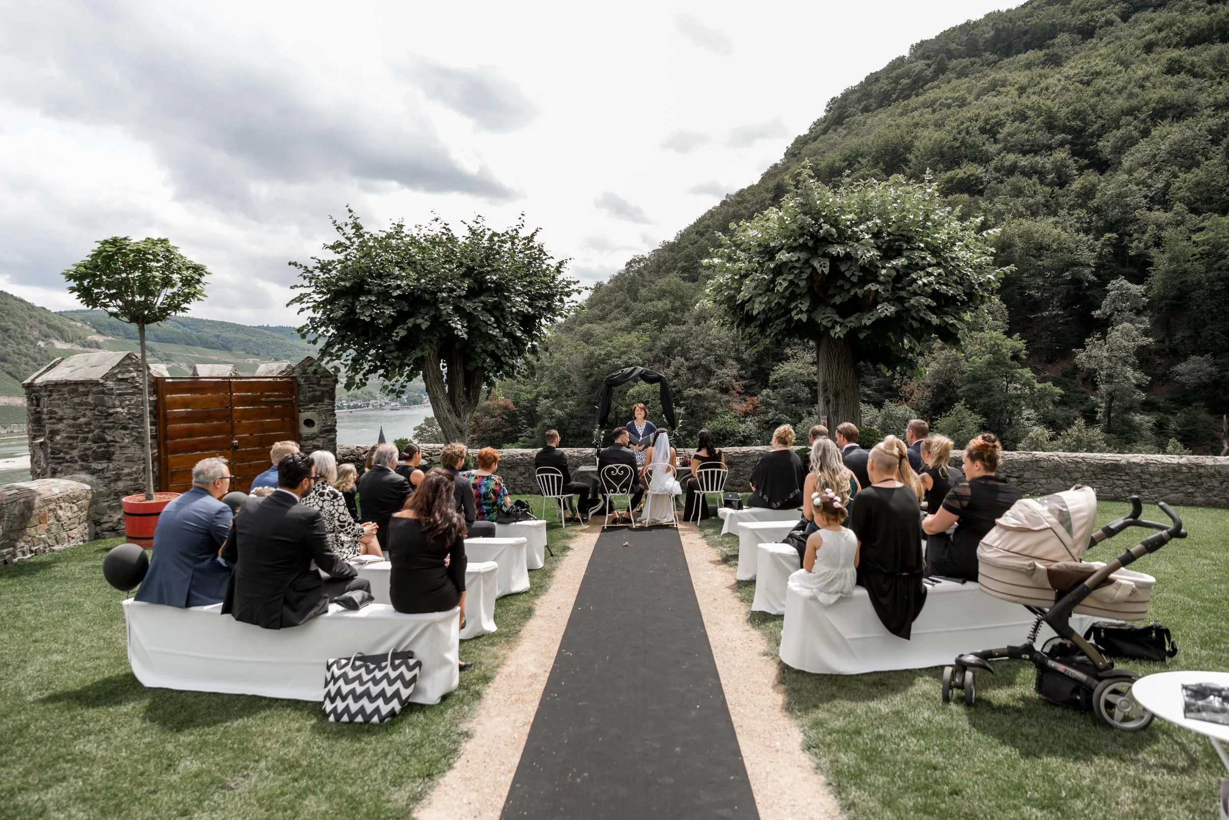 Outdoor wedding ceremony with guests seated on white benches along a black aisle runner, under large trees, overlooking a river and hillside, during daytime with cloudy sky.