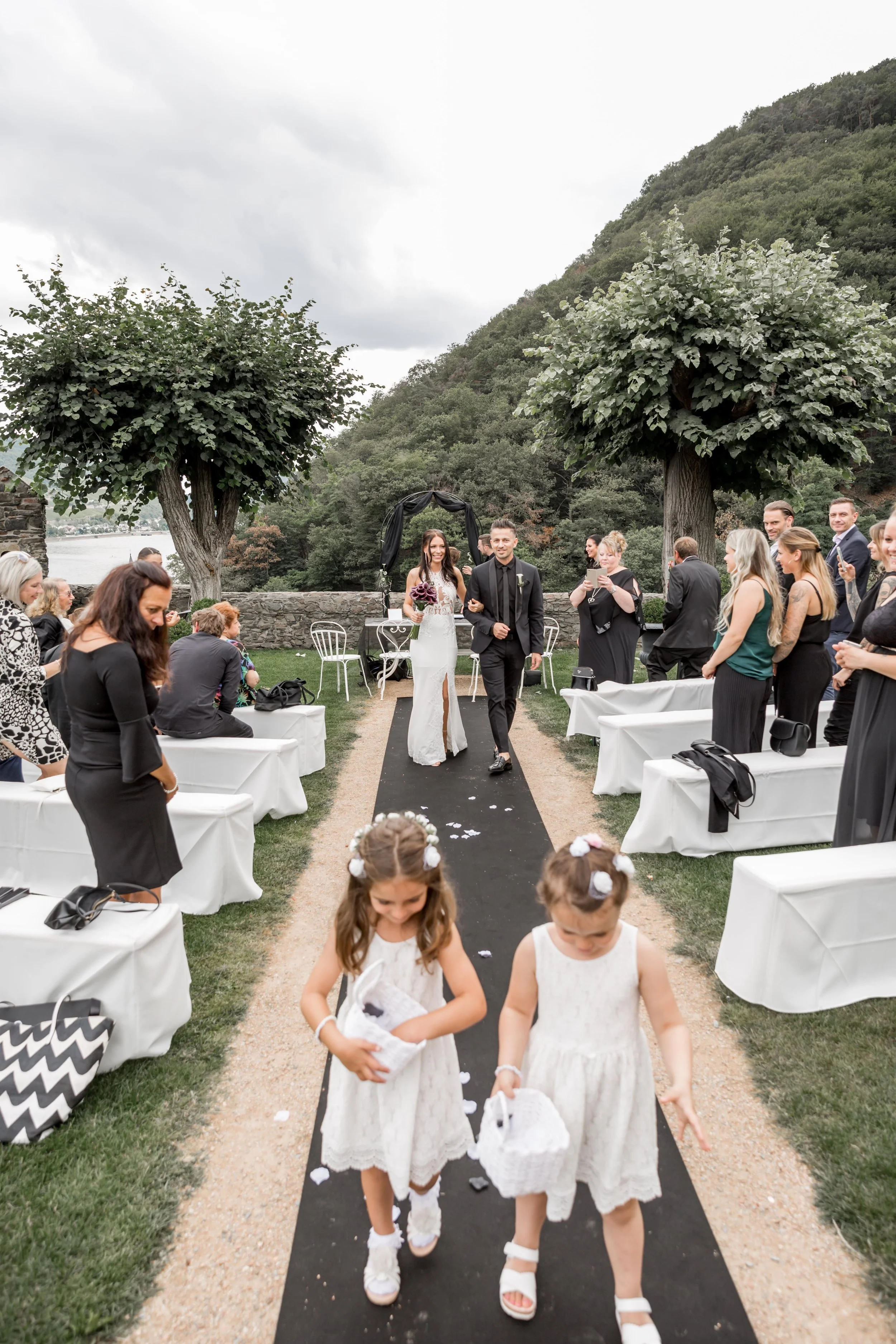 A wedding ceremony outdoors on a grassy area with trees and hills in the background. A bride in a white gown and a groom in a dark suit walk down the aisle, flanked by seated guests. Two young girls in white dresses walk ahead holding baskets.