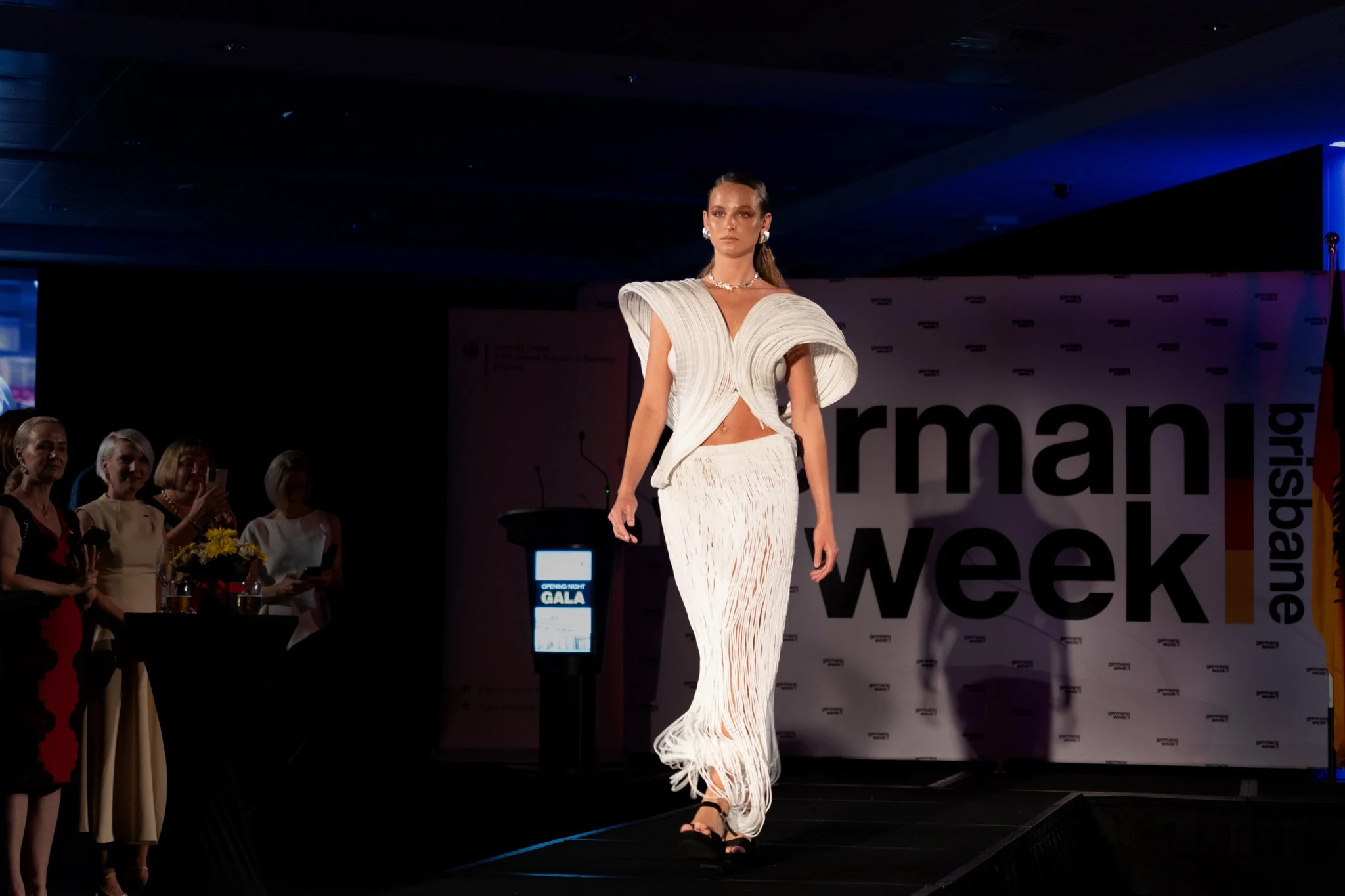 A female model walks down the runway at a fashion show during German Fashion Week, wearing an elegant white dress with intricate design and accessories.