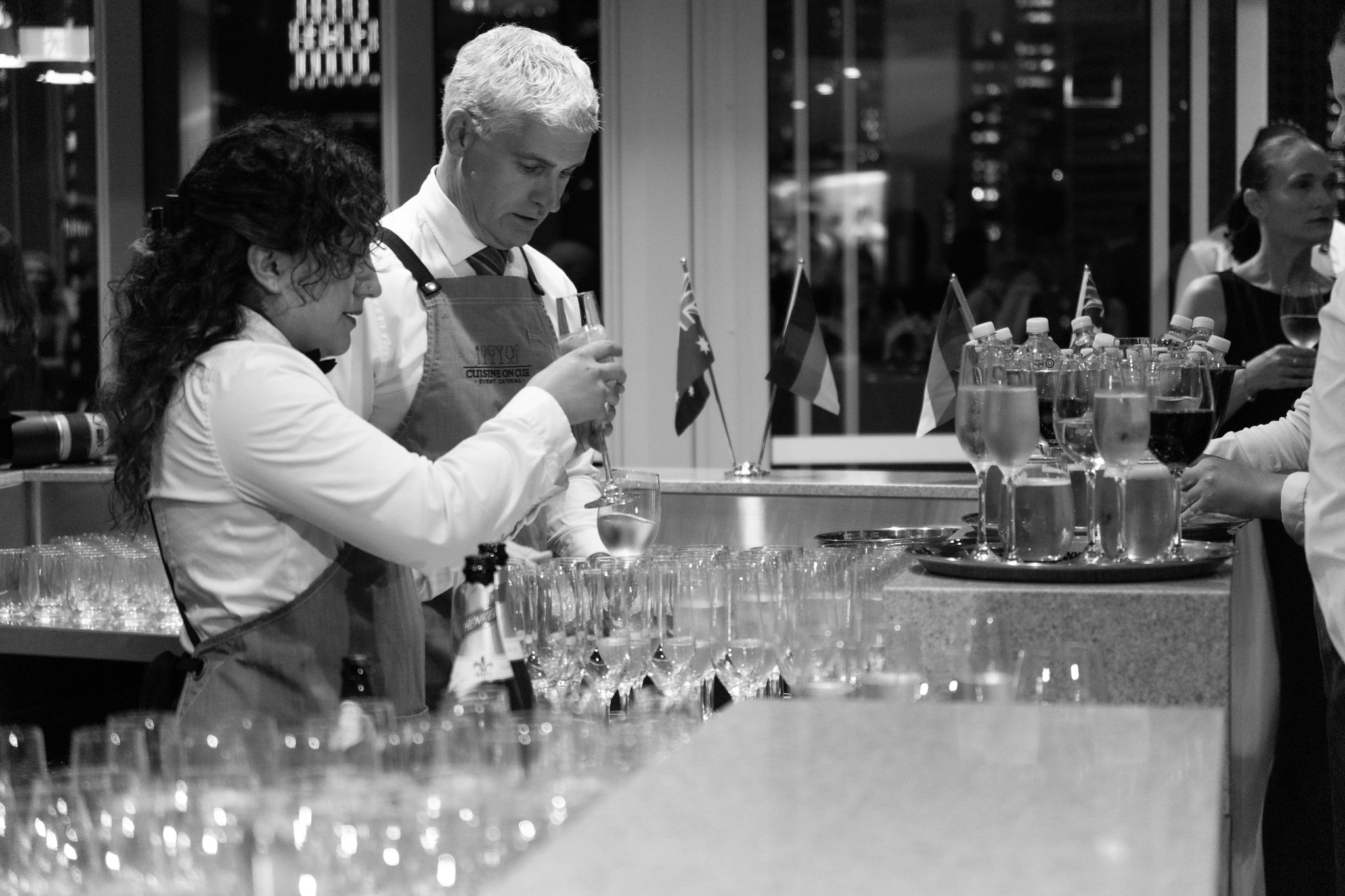 Two bartenders preparing drinks at a bar counter in a restaurant or bar setting, with glasses and flags displayed in the background.