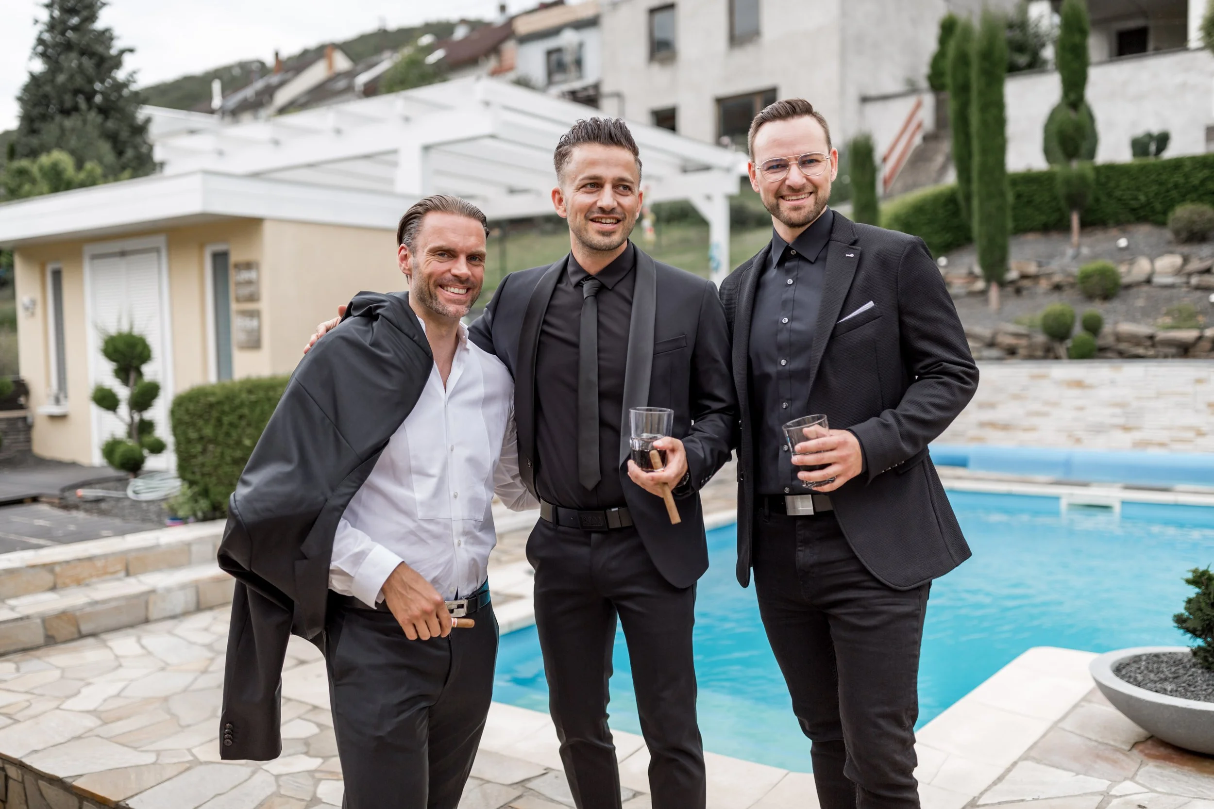 Three men in suits and a white shirt smiling and holding drinks by a swimming pool outdoors.