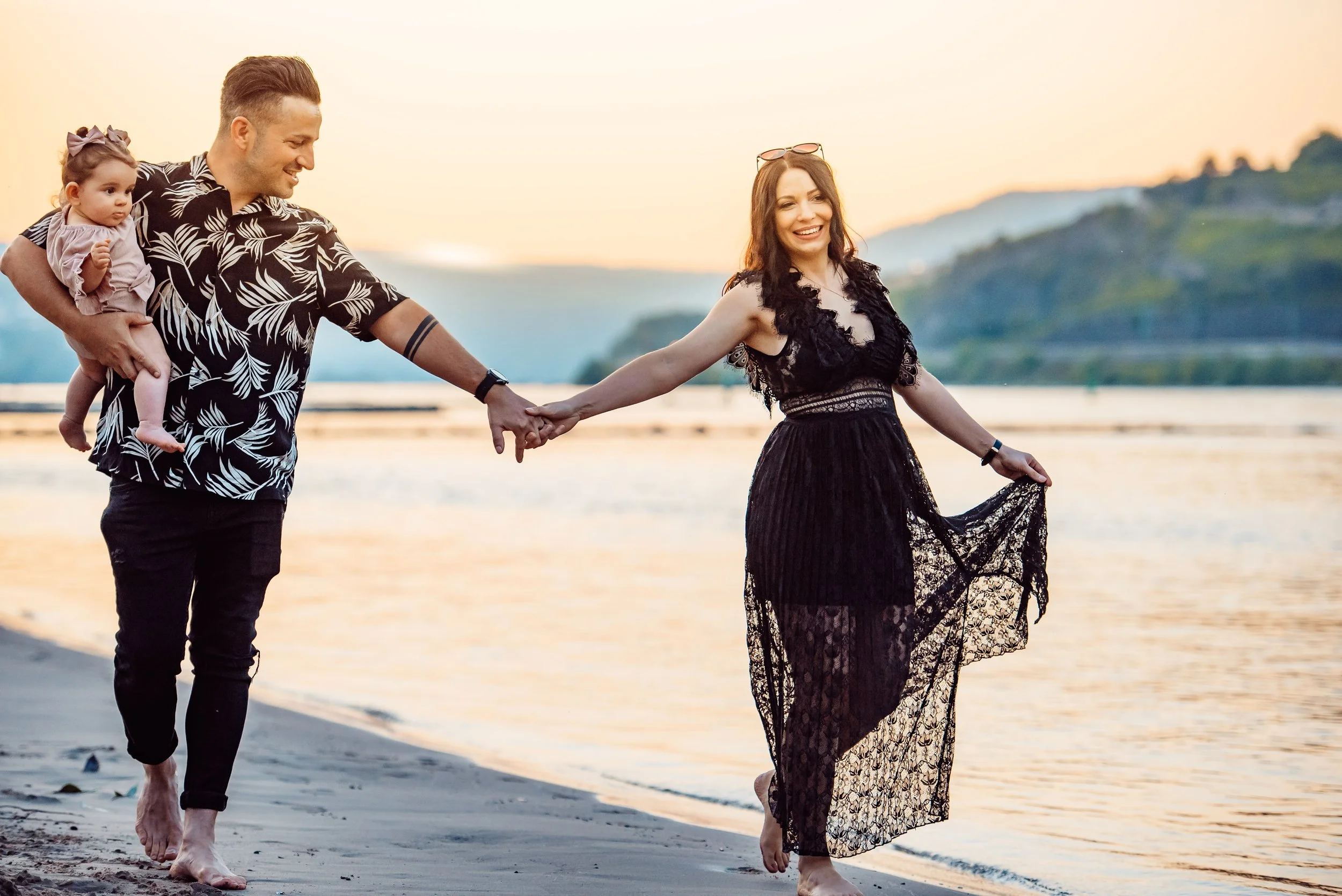 A family of three walking along a beach at sunset, holding hands and smiling. The father is carrying a young girl in his arm, and the mother is wearing a black lacy dress.