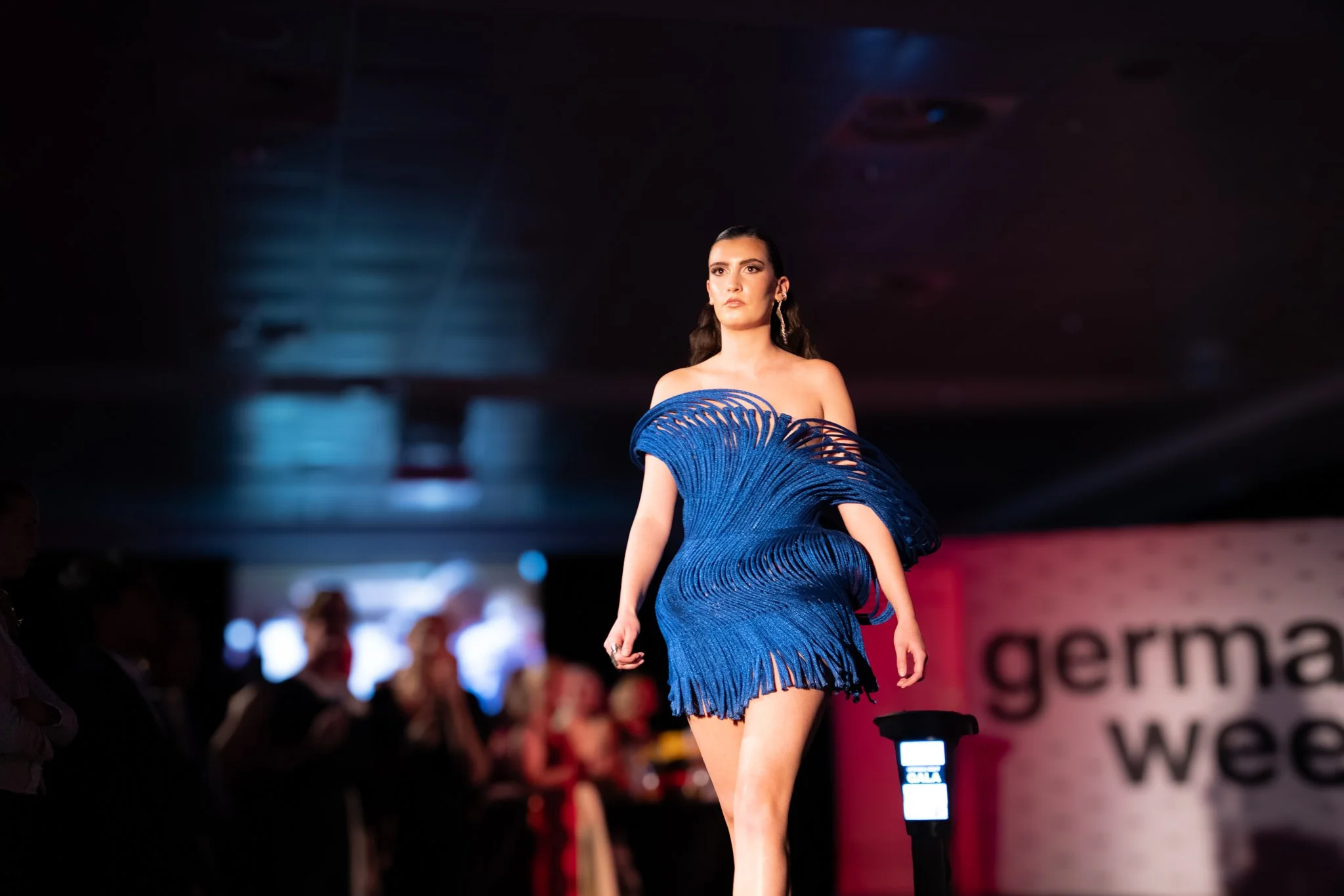 Fashion model walking down runway wearing a striking blue dress with intricate cut-out details and fringes, during a fashion show.