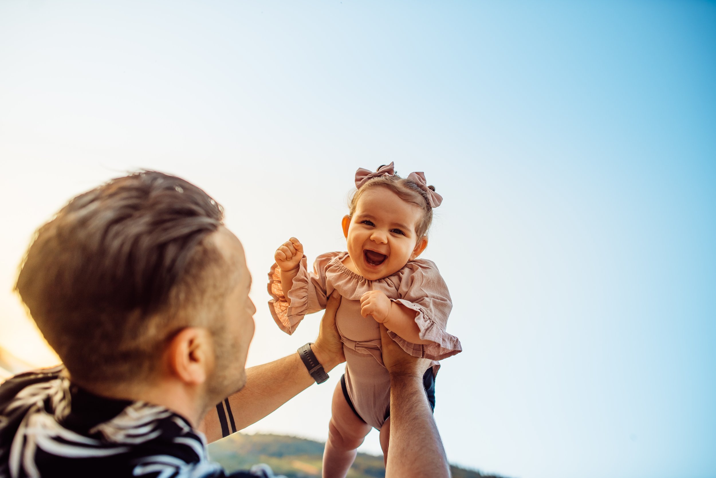 A man lifting a smiling baby girl against a bright sky.