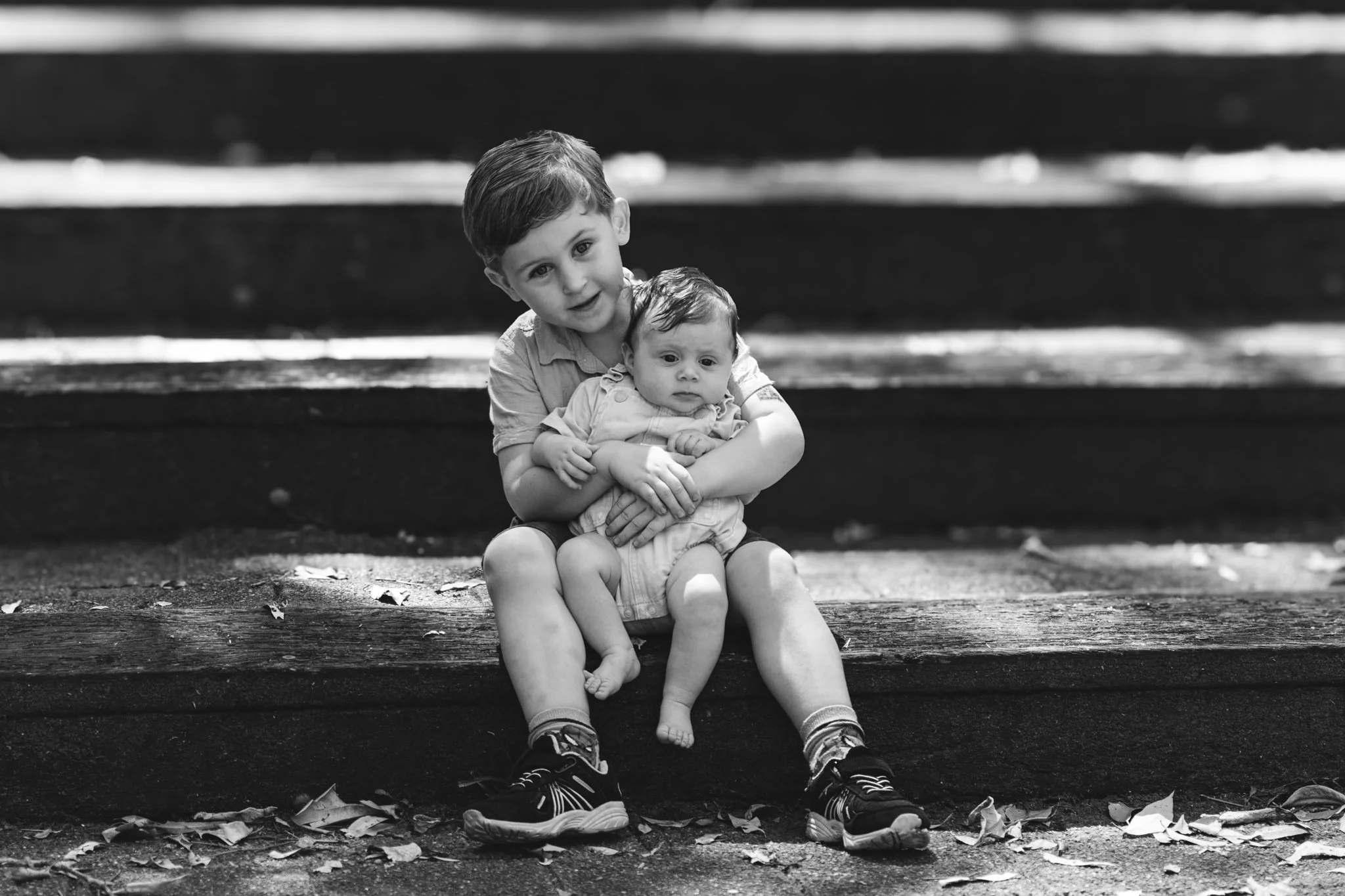 Two young children sitting on outdoor wooden steps, one older boy holding a younger baby in his lap, black and white photo.