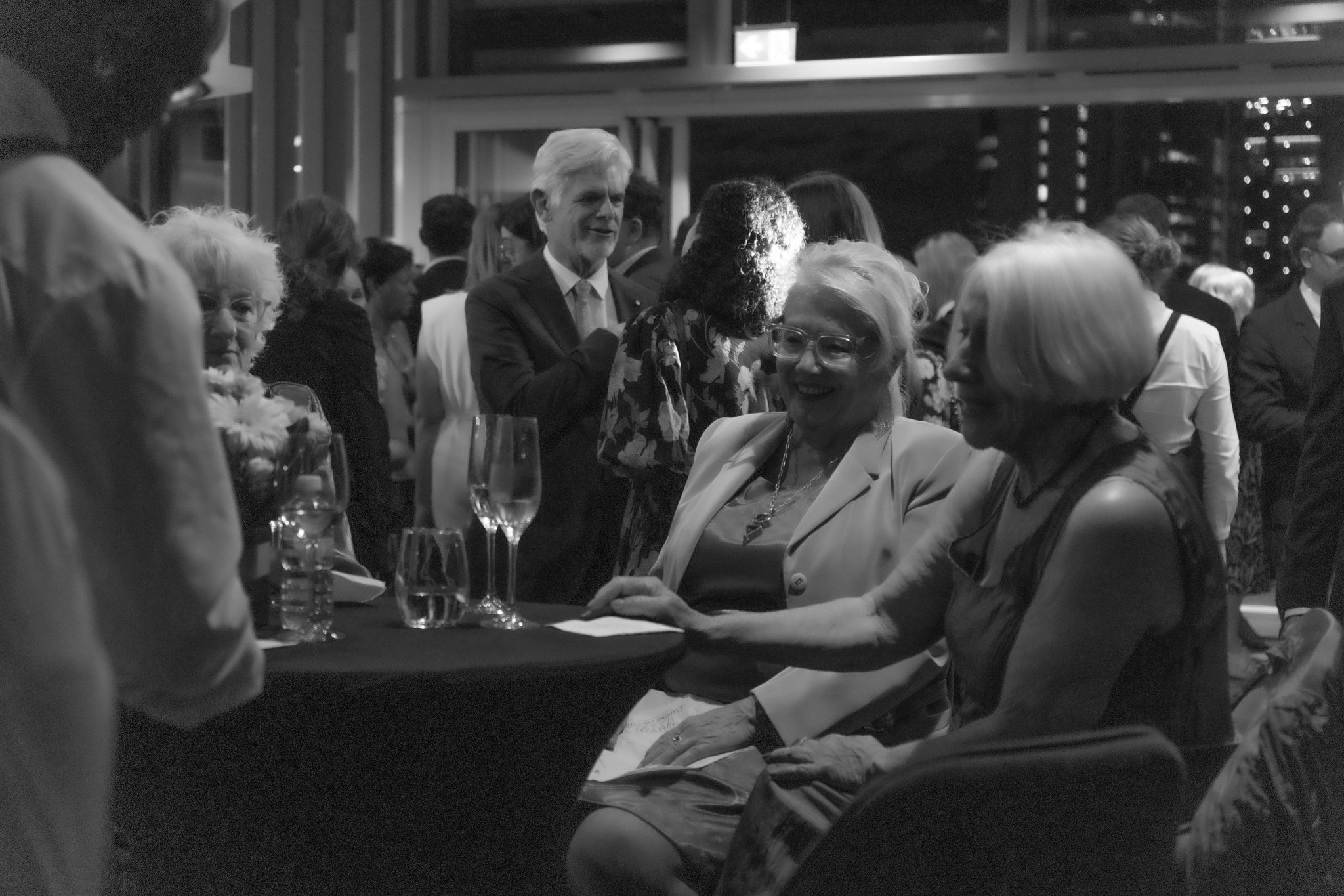 A black and white photo of a social gathering with people talking and smiling, indoors with large windows and city lights outside.