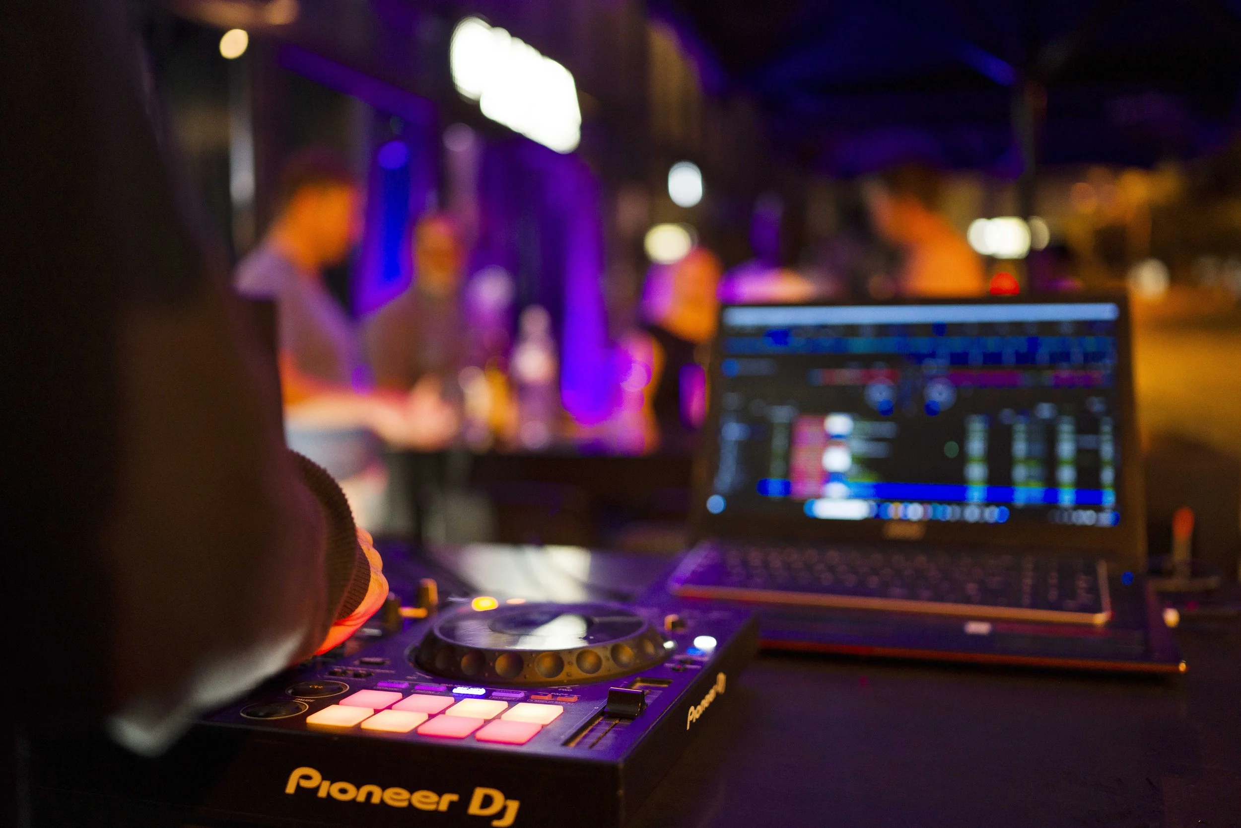 Close-up of DJ equipment, including a Pioneer DJ controller and a laptop with DJ software, at a nightclub with blurred people in the background under colorful lighting.