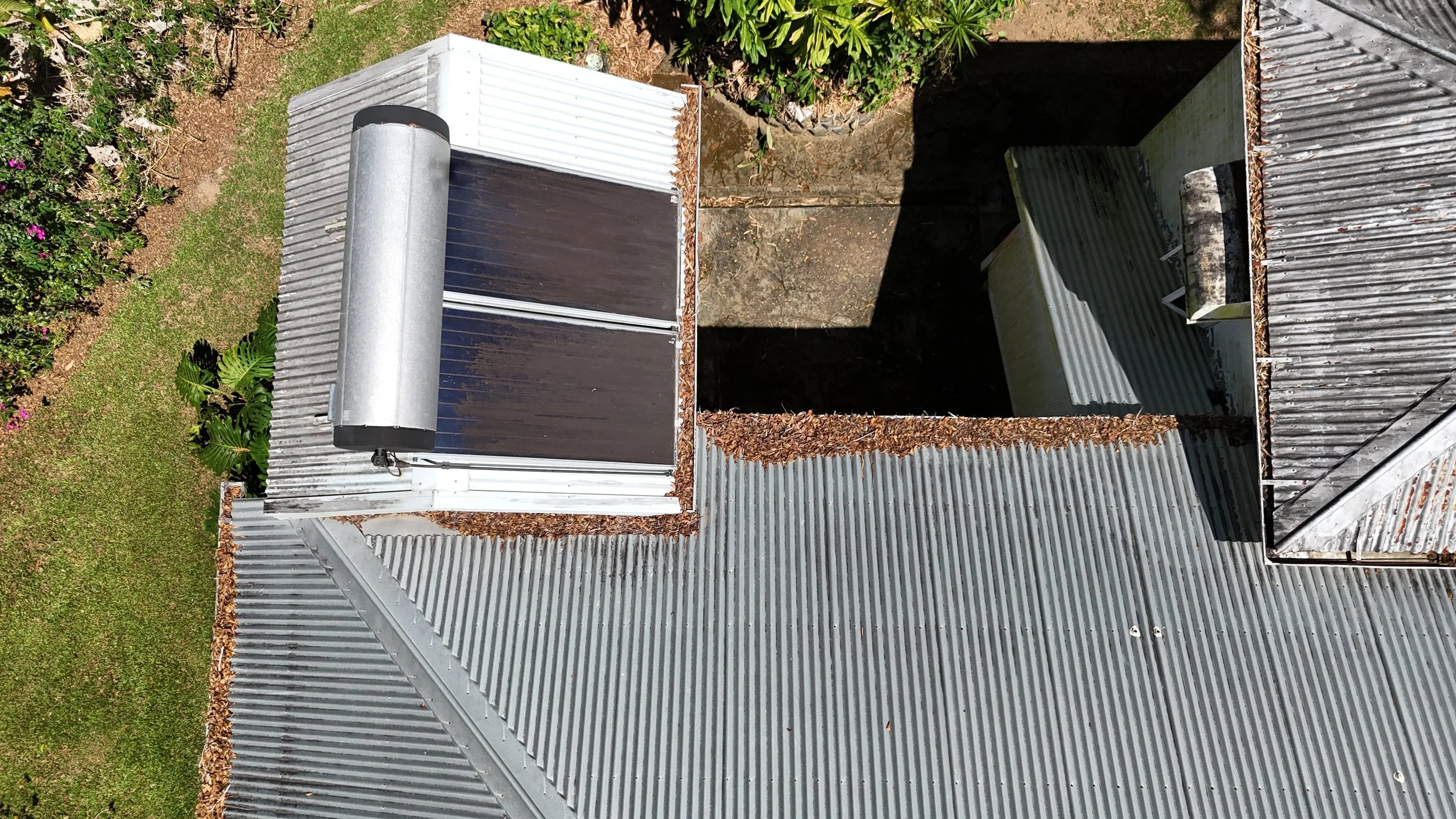 A view looking down at a small rooftop structure with a cylindrical vent, surrounding gray metal roofing, and an adjacent open area with leaves and debris. Vegetation and grass are visible nearby.